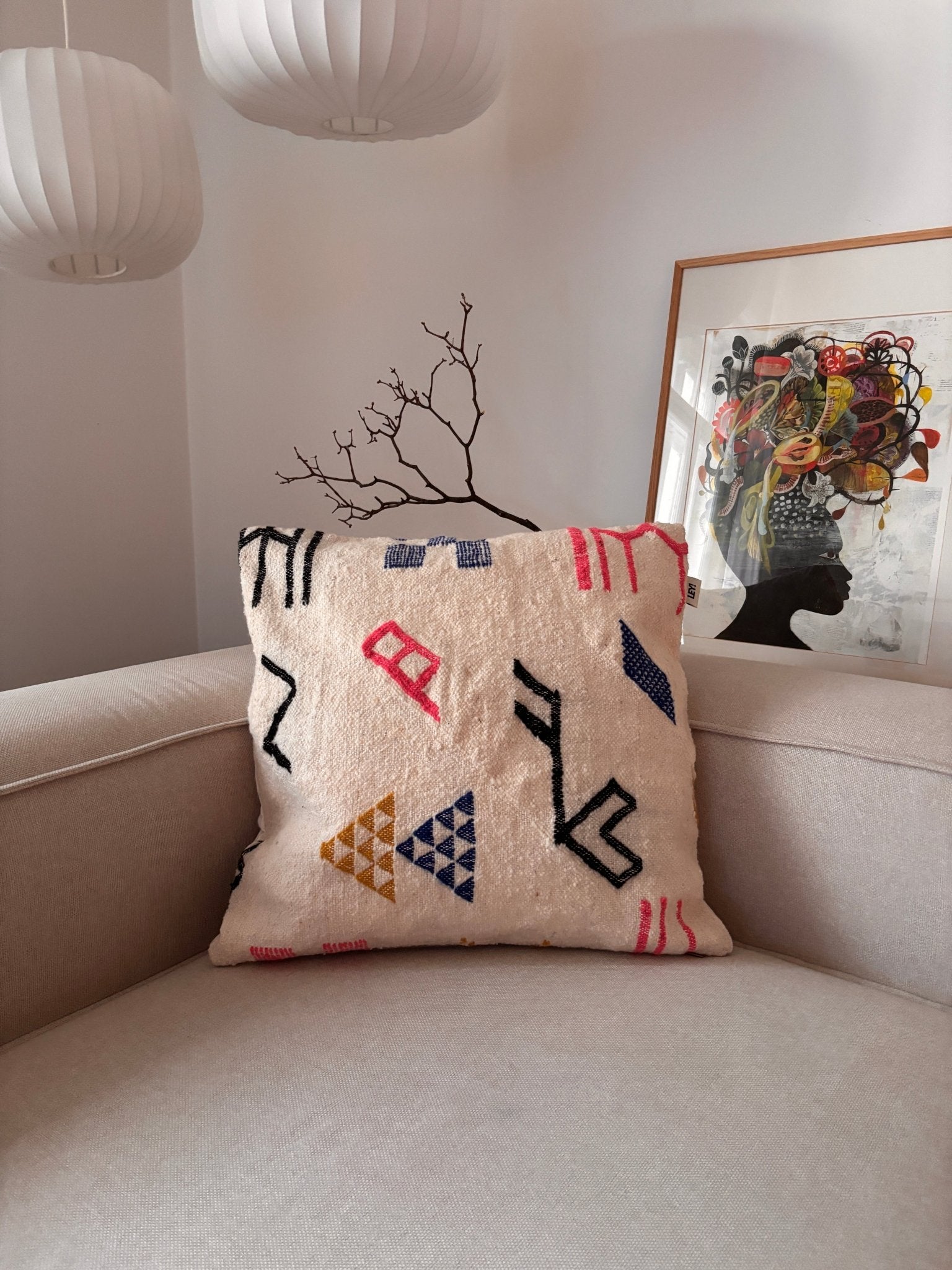 Ein LEY!-Kissen – Colorful Line – 60 x 60 liegt auf einem hellen Sofa, im Hintergrund sind Kunstwerke und Lampen zu sehen.