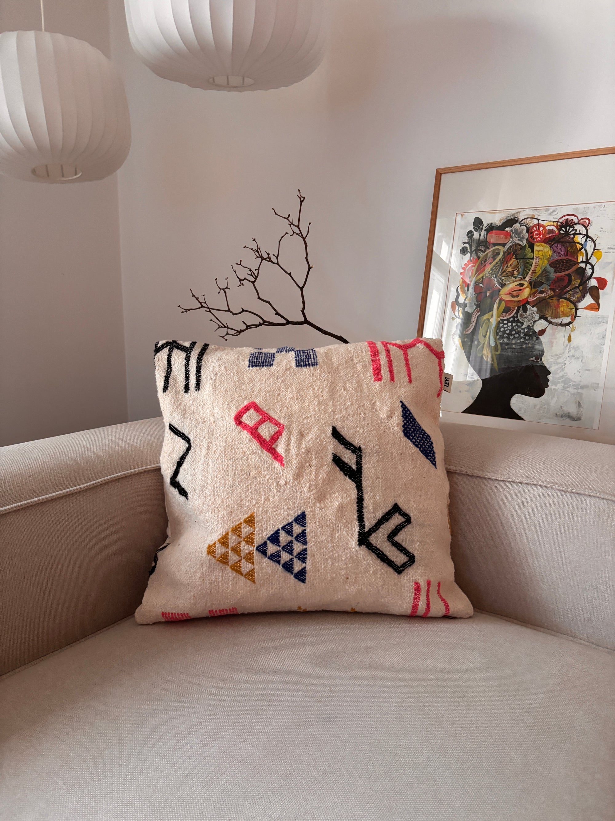 Ein LEY!-Kissen – Colorful Line – 60 x 60 liegt auf einem hellen Sofa, im Hintergrund sind Kunstwerke und Lampen zu sehen.