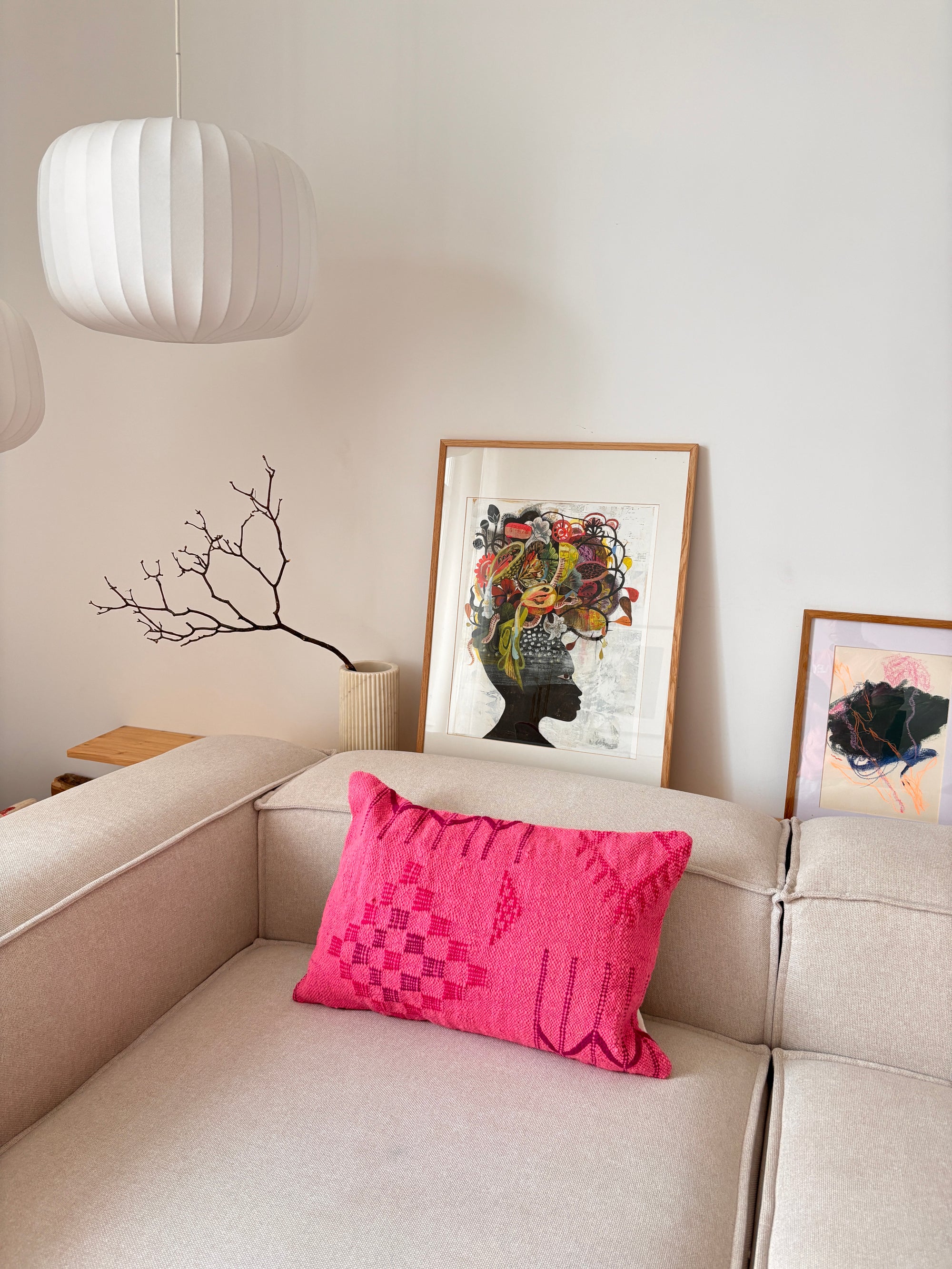 Ein beiges Sofa mit einem Kissen aus der „LEY! Colorful Line PINK EDITION“, gerahmte Kunstwerke und eine weiße Pendelleuchte in einem modernen Raum.