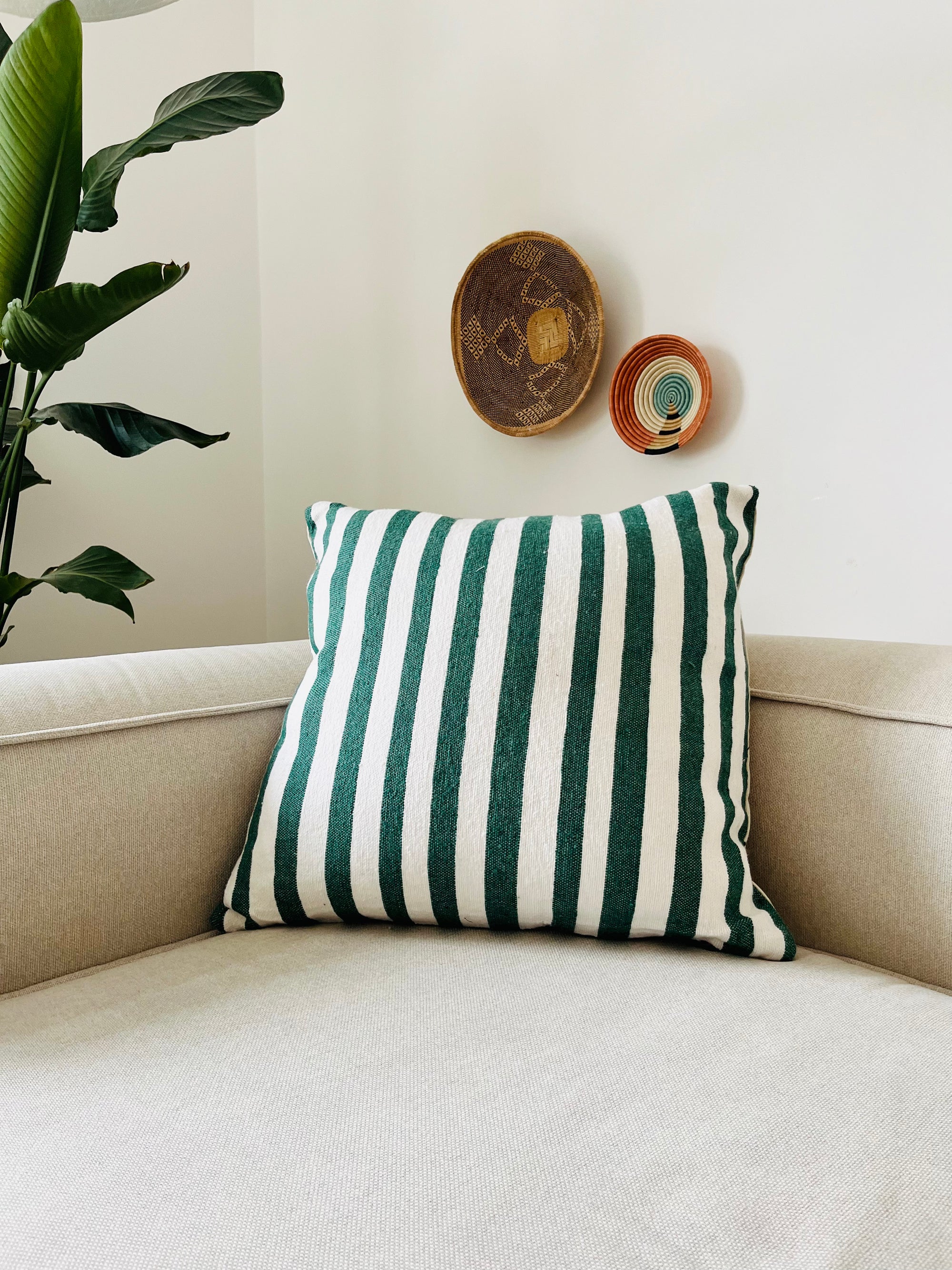 Ein beiges Sofa mit einem grün-weißen Kissen - Tilosa 60x60 von LEY! sitzt in der Ecke. Darüber hängen drei handgefertigte geflochtene Marrakesch-Körbe, und links steht eine große Blattpflanze.