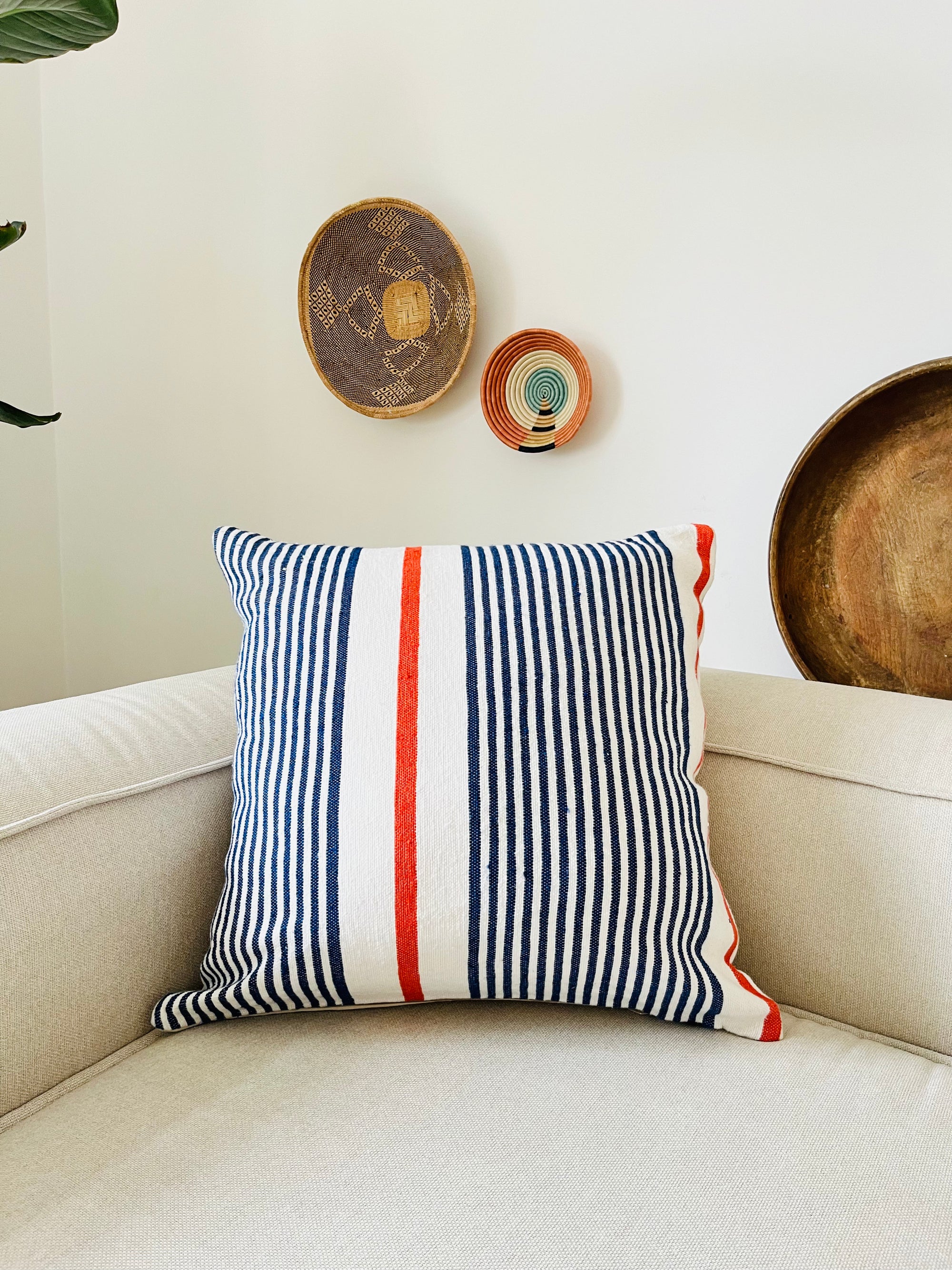 Ein beiges Sofa mit einem quadratischen LEY! Kissen - Parosa 60x60, mit vertikalen Streifen in Marineblau, Weiß und Rot. Dekorative geflochtene Körbe und Teller sind an der weißen Wand hinter dem Sofa angebracht.
