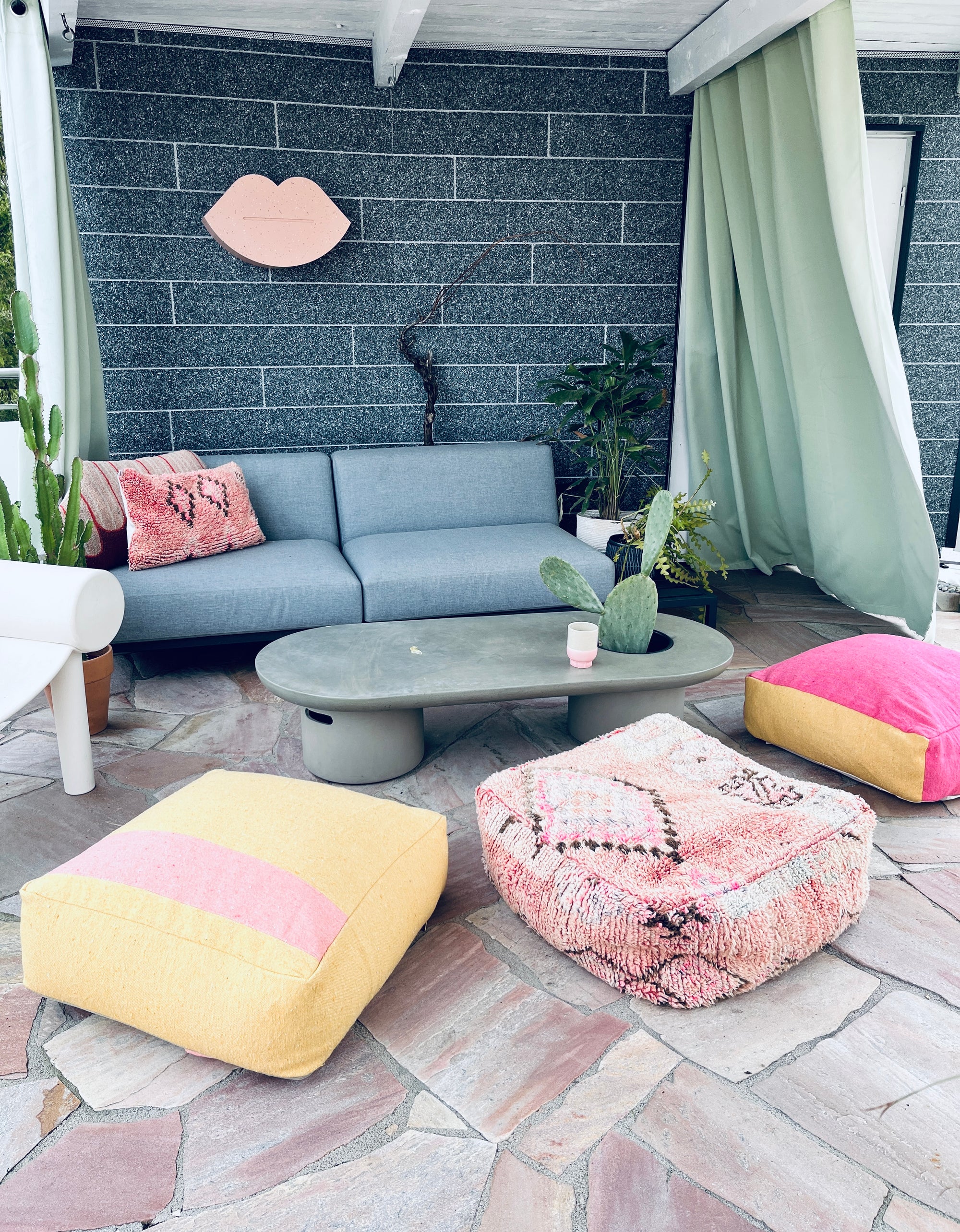 Ein moderner Innenhof mit einem grauen Sofa, einem grünen Couchtisch, Topfpflanzen und bunten Bodenpolstern. Darüber hängt eine Wanddekoration mit rosa Lippen, und grüne Vorhänge verdecken den Raum teilweise.