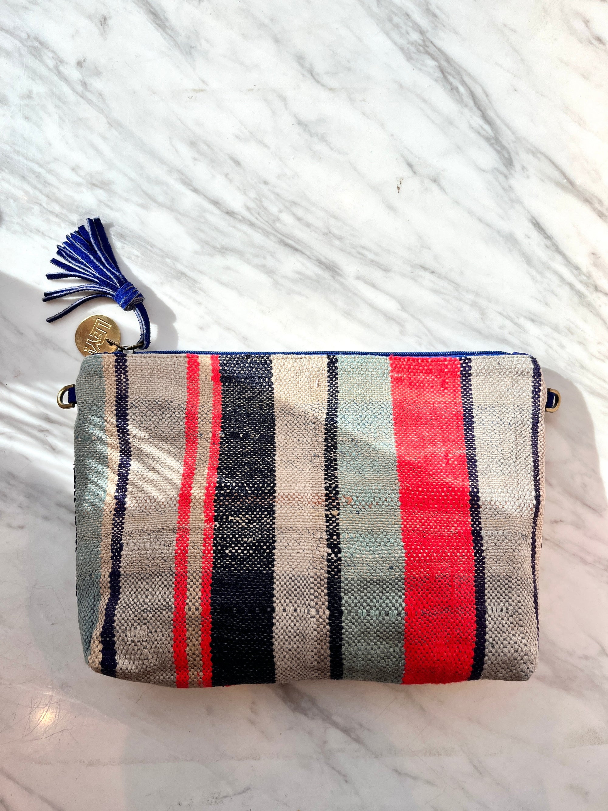 Die TASCHE von LEY! ist eine handgenähte Clutch mit blauen, roten und beigen Streifen, blauem Reißverschluss und Quaste mit Gold-Charme-ein Einzelstück, präsentiert auf weißem Marmor.