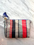 Die TASCHE von LEY! ist eine handgenähte Clutch mit blauen, roten und beigen Streifen, blauem Reißverschluss und Quaste mit Gold-Charme-ein Einzelstück, präsentiert auf weißem Marmor.