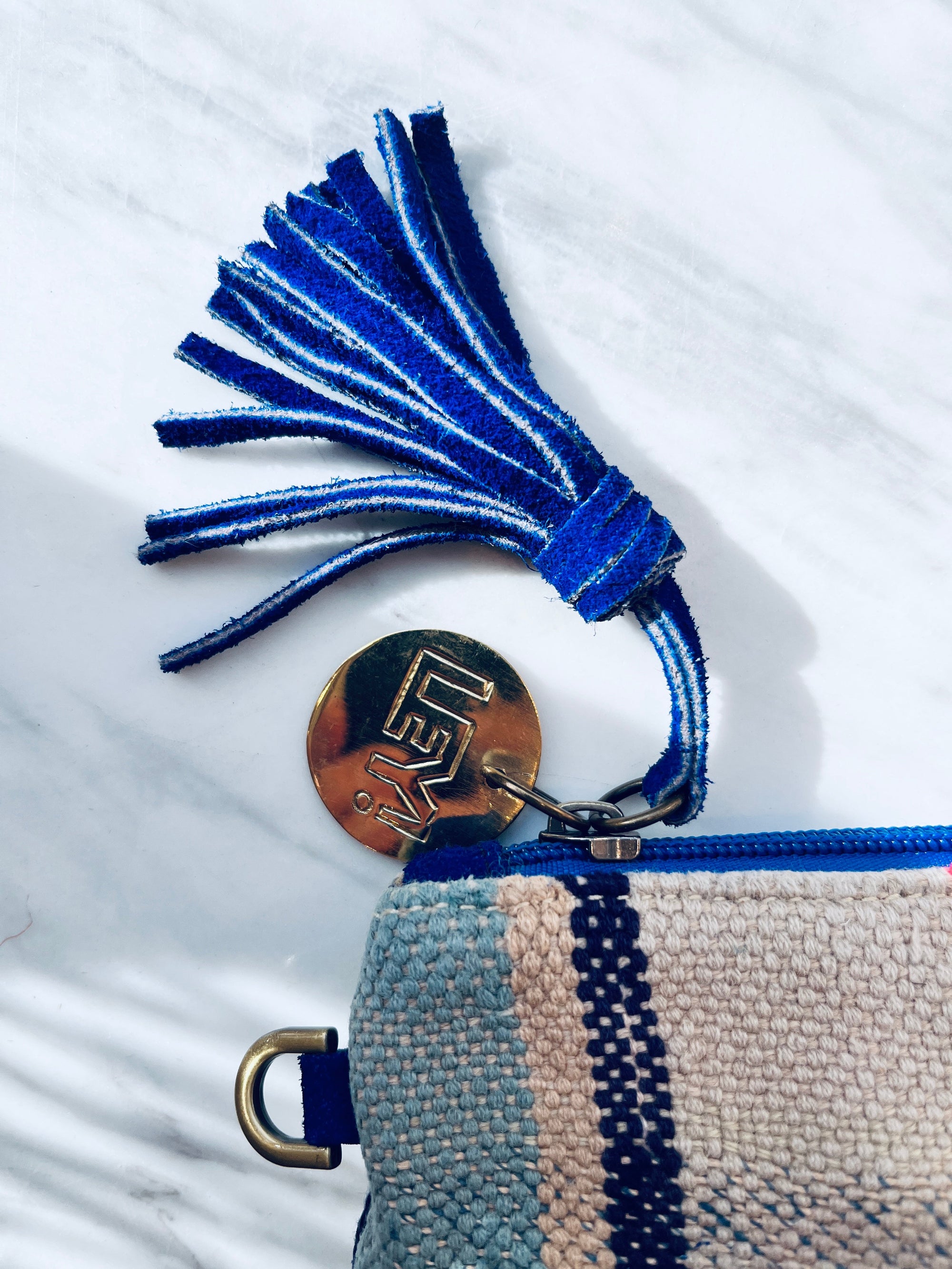 Eine Nahaufnahme der LEY! TASCHE - eine blau-beige gewebte Clutch mit blauem Reißverschluss, Wildlederquaste und einem runden goldenen Schlüsselanhänger mit geometrischen Buchstaben - ein elegantes Einzelstück auf weißem Marmor.