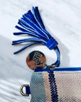 Eine Nahaufnahme der LEY! TASCHE - eine blau-beige gewebte Clutch mit blauem Reißverschluss, Wildlederquaste und einem runden goldenen Schlüsselanhänger mit geometrischen Buchstaben - ein elegantes Einzelstück auf weißem Marmor.