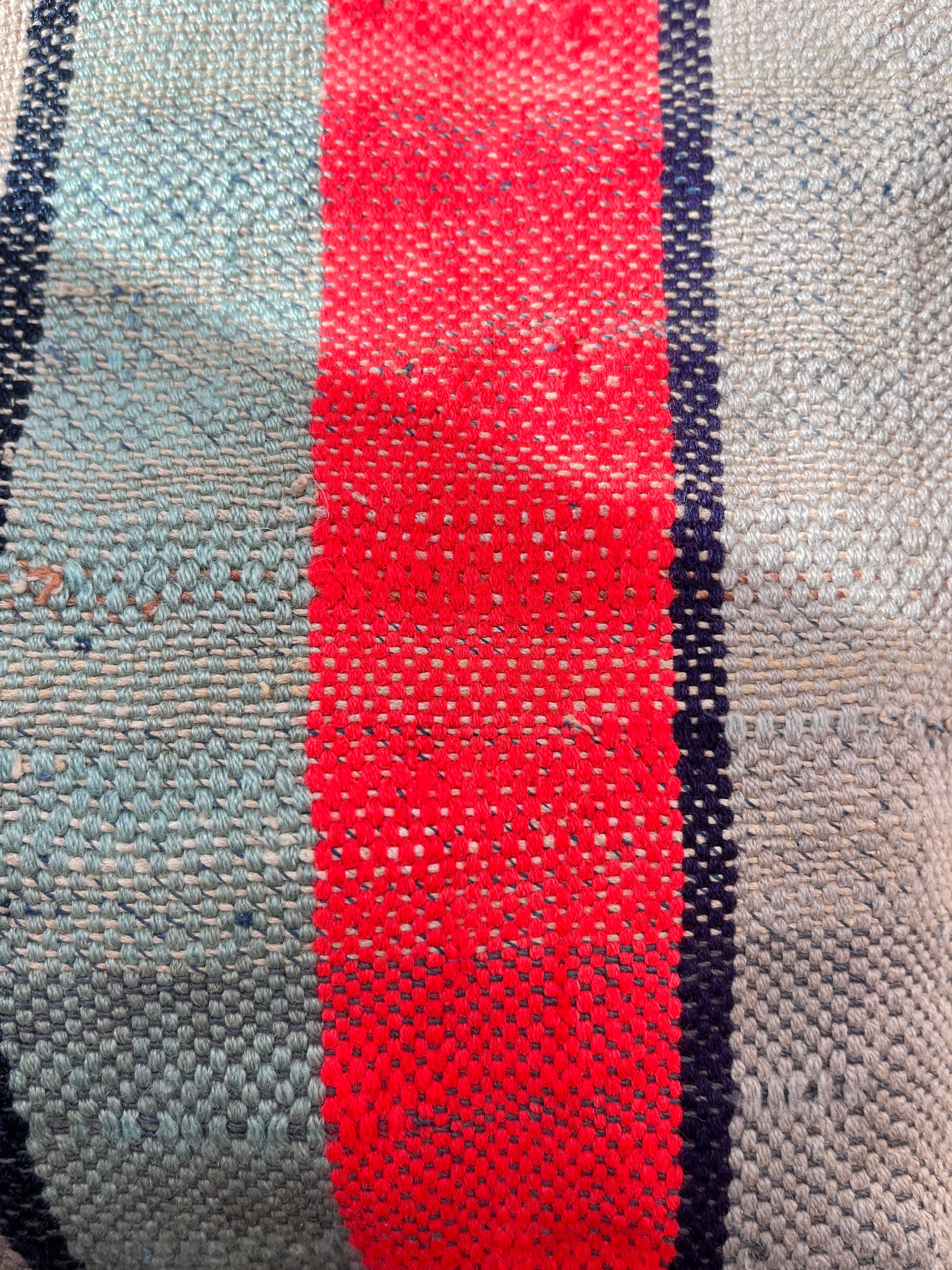 Eine Nahaufnahme des gewebten Stoffes auf dem LEY! TASCHE Einzelstück zeigt einen leuchtend roten vertikalen Streifen, der von dünnen dunkelblauen Linien begrenzt wird, mit hellblauen und grauen Abschnitten. Die Textur und die einzelnen Fäden sind deutlich zu erkennen.