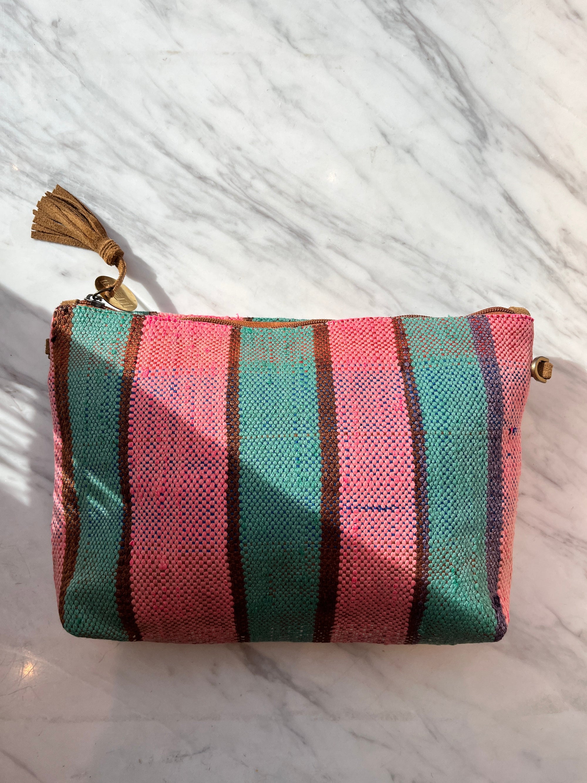 Die LEY! TASCHE ist eine kleine Stofftasche mit vertikalen rosa und blaugrünen Streifen, dünnen braunen Linien und einer braunen Quaste am Reißverschluss. Dieses einzigartige Stück wird auf einer weißen Marmoroberfläche im Sonnenlicht gezeigt.
