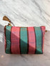Die LEY! TASCHE ist eine kleine Stofftasche mit vertikalen rosa und blaugrünen Streifen, dünnen braunen Linien und einer braunen Quaste am Reißverschluss. Dieses einzigartige Stück wird auf einer weißen Marmoroberfläche im Sonnenlicht gezeigt.