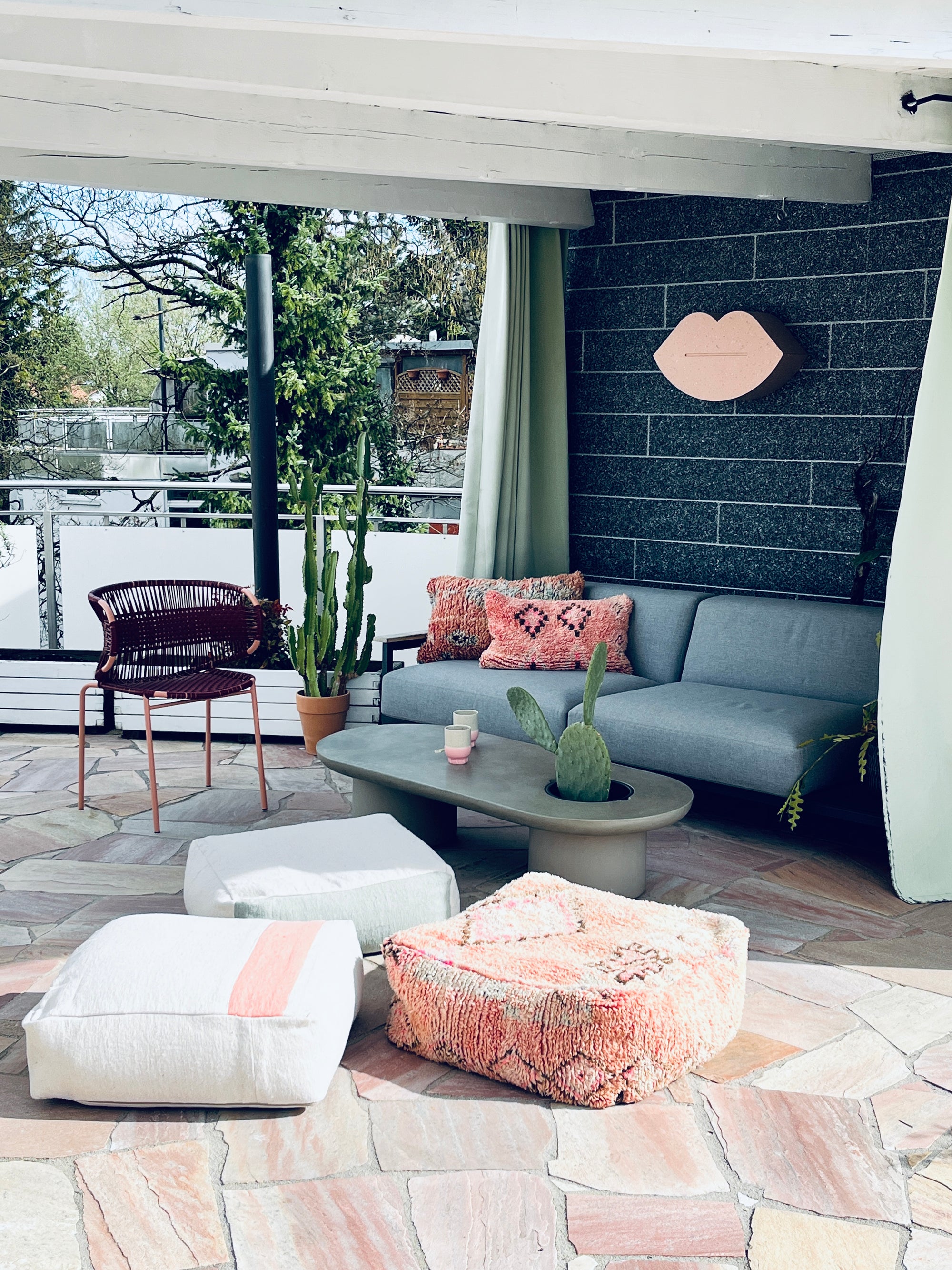 Eine stilvolle Außenterrasse mit einem grauen Sektionssofa, rosa und weiß gemusterten Kissen, getopften Kakteen, einem runden Couchtisch, zwei Poufs, einem geflochtenen Stuhl und einem Wandgemälde mit rosa Lippen an einer dunklen Ziegelwand.