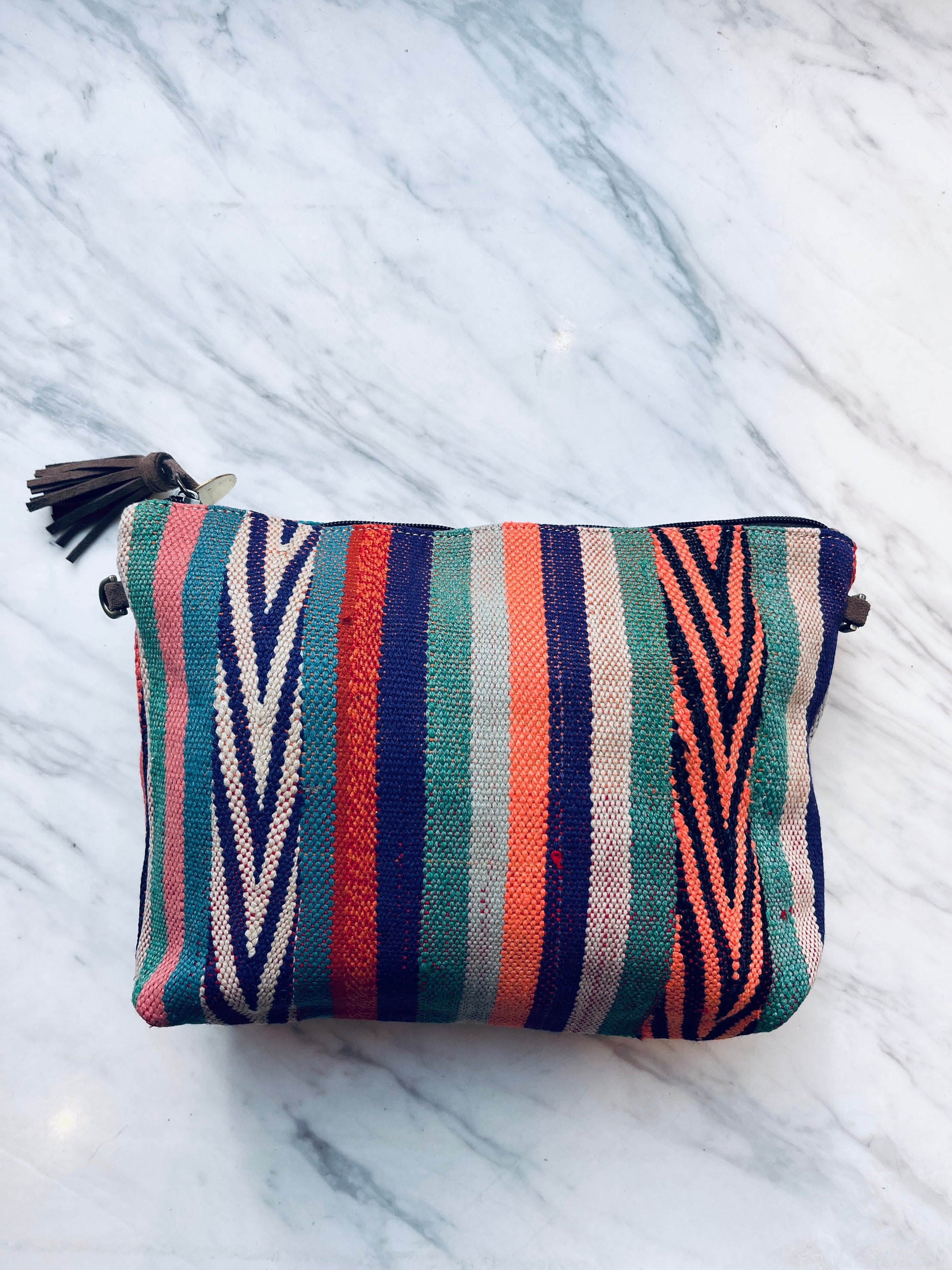 Eine lebendige, handgenähte TASCHE Tasche von LEY! mit einem Reißverschluss, vertikalen grünen, blauen, roten, orange-weißen Streifen und einer braunen Quaste liegt anmutig auf einer Marmoroberfläche.