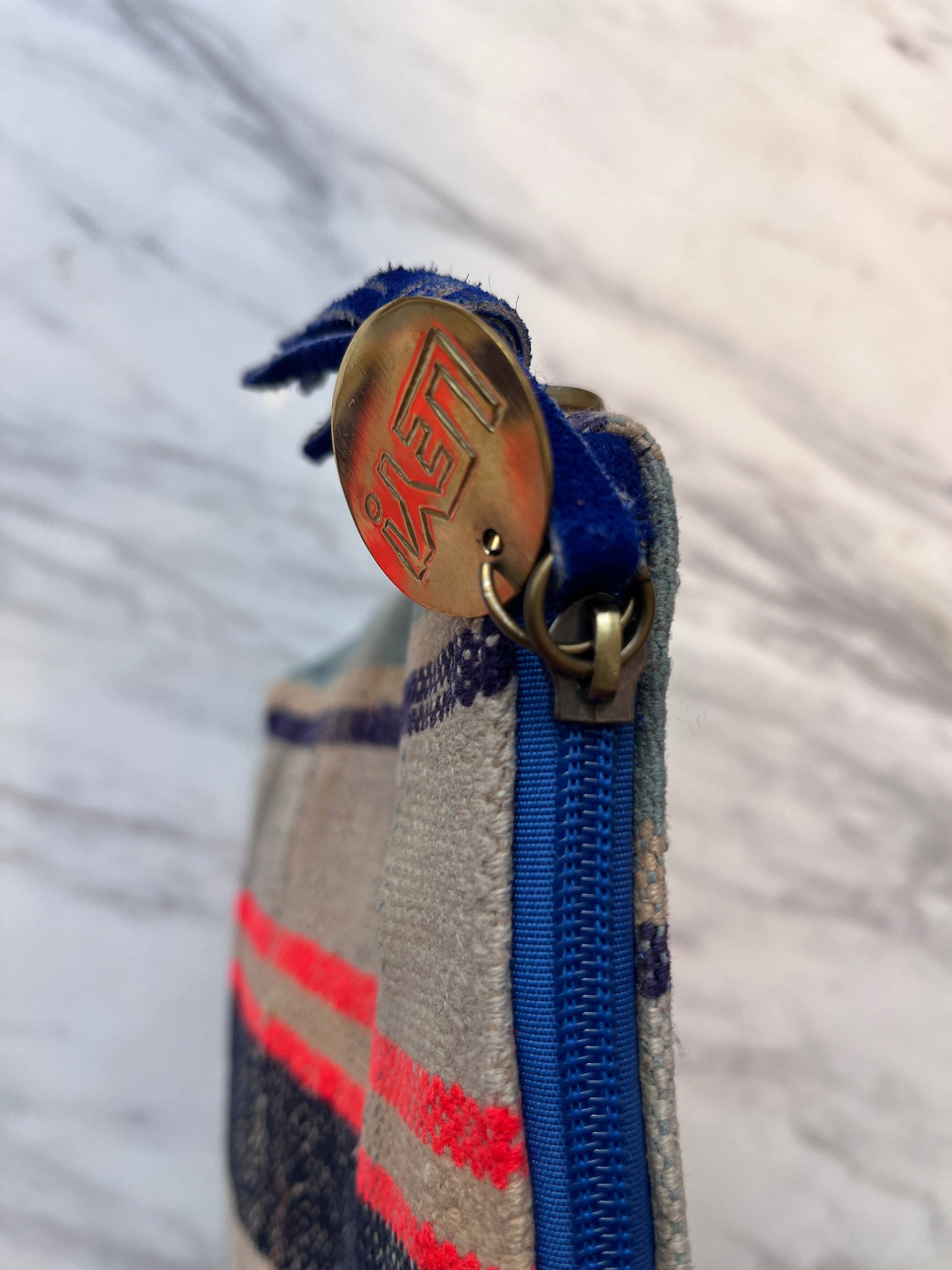 Nahaufnahme der Stoff-Clutch TASCHE von LEY! mit blauem Reißverschluss und runder Metalllasche mit der Gravur "LEY!" sowie handgestickten Taschendetails in roten, blauen und beigen Streifen vor einem marmorierten Hintergrund.