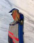 Nahaufnahme der Stoff-Clutch TASCHE von LEY! mit blauem Reißverschluss und runder Metalllasche mit der Gravur "LEY!" sowie handgestickten Taschendetails in roten, blauen und beigen Streifen vor einem marmorierten Hintergrund.