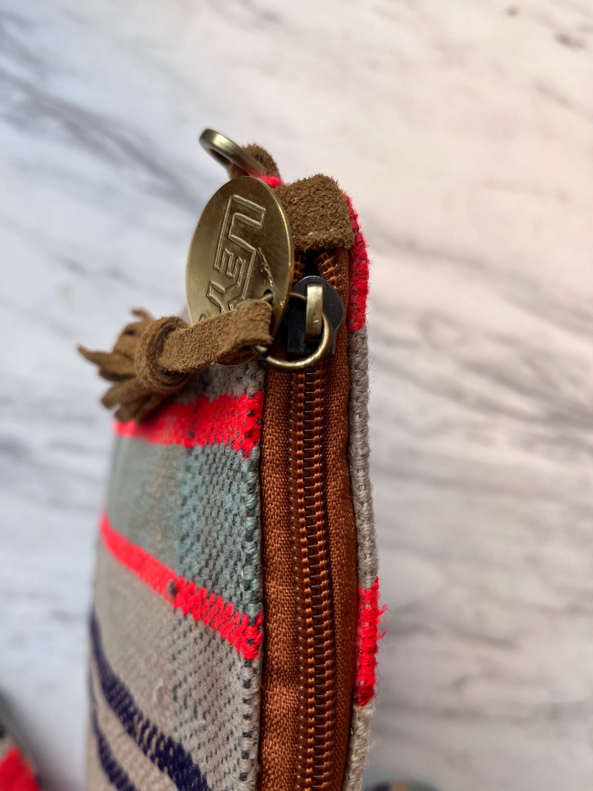 Nahaufnahme der TASCHE by LEY! - eine einzigartige, handgenähte Tasche mit roten und blauen Streifen, Lederakzenten, brauner Wildlederquaste und einem Messing-Reißverschluss mit eingraviertem Markennamen - auf einer weiß marmorierten Oberfläche.