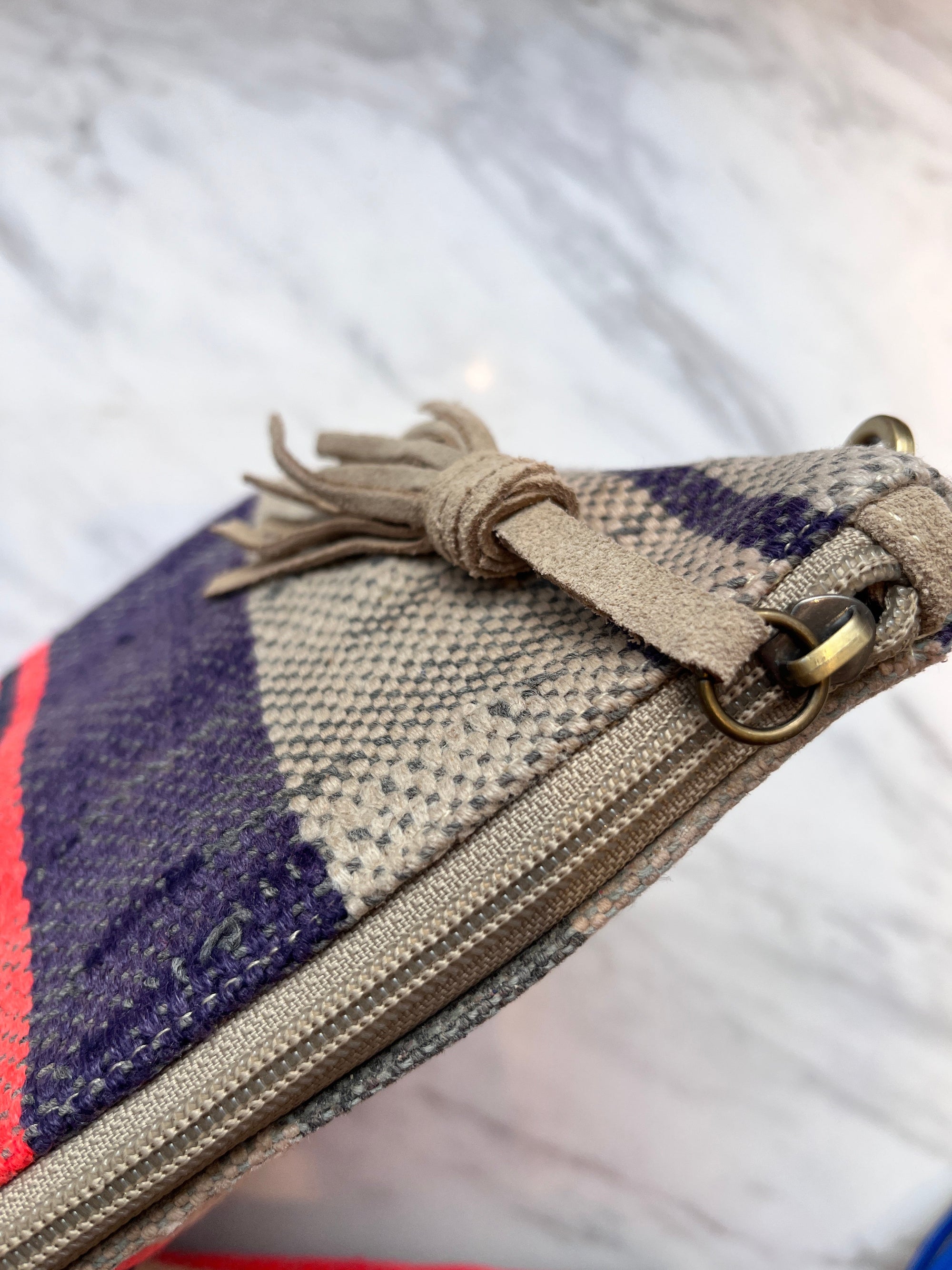 Nahaufnahme einer handgenähten LEY! TASCHE Clutch mit beigefarbenem Reißverschluss und Quaste, die mit einem Messingring versehen ist. Dieses Einzelstück zeigt beige, lila und rote Streifen auf einem marmorierten weißen Hintergrund.