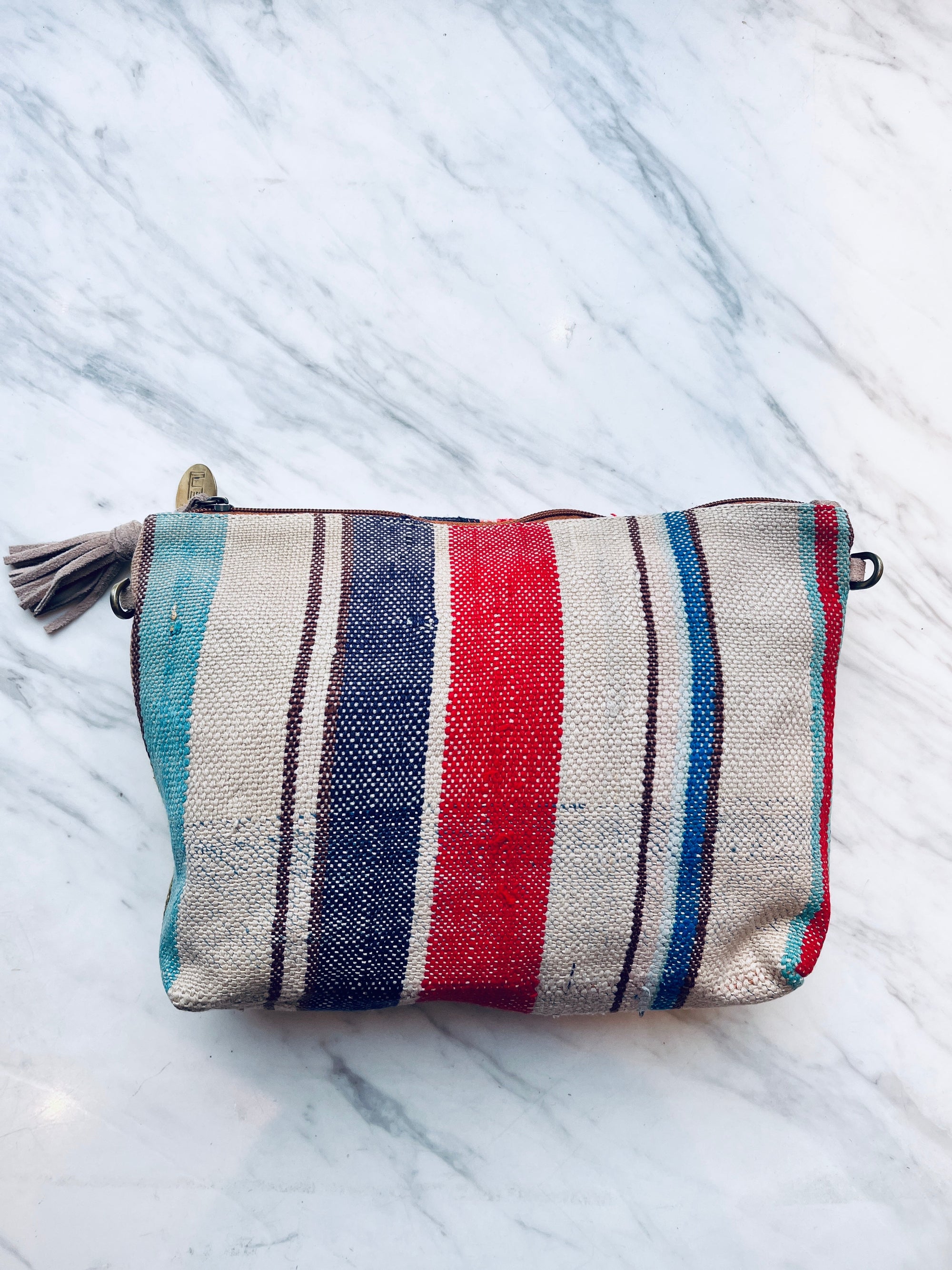 Die LEY! TASCHE ist eine rechteckige, gewebte Clutch mit blauen, roten, türkisfarbenen und braunen Längsstreifen und einem Reißverschluss mit Quasten, der auf Marmor ausgestellt ist. Dieses Einzelstück bringt einzigartiges Flair in Ihre handgenähte Taschensammlung.