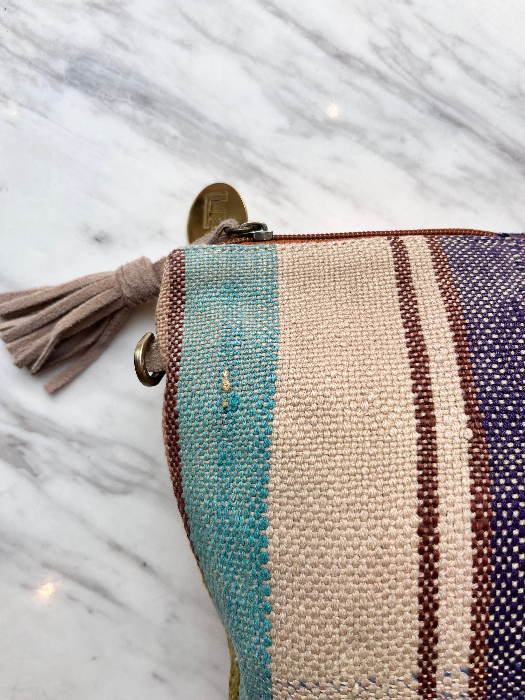 Eine Nahaufnahme der LEY! TASCHE Clutch mit Reißverschluss und beiger Quaste auf weißem Marmor, mit gewebten türkisfarbenen, beigen, burgunderroten und lila Streifen für einen einzigartigen Look.