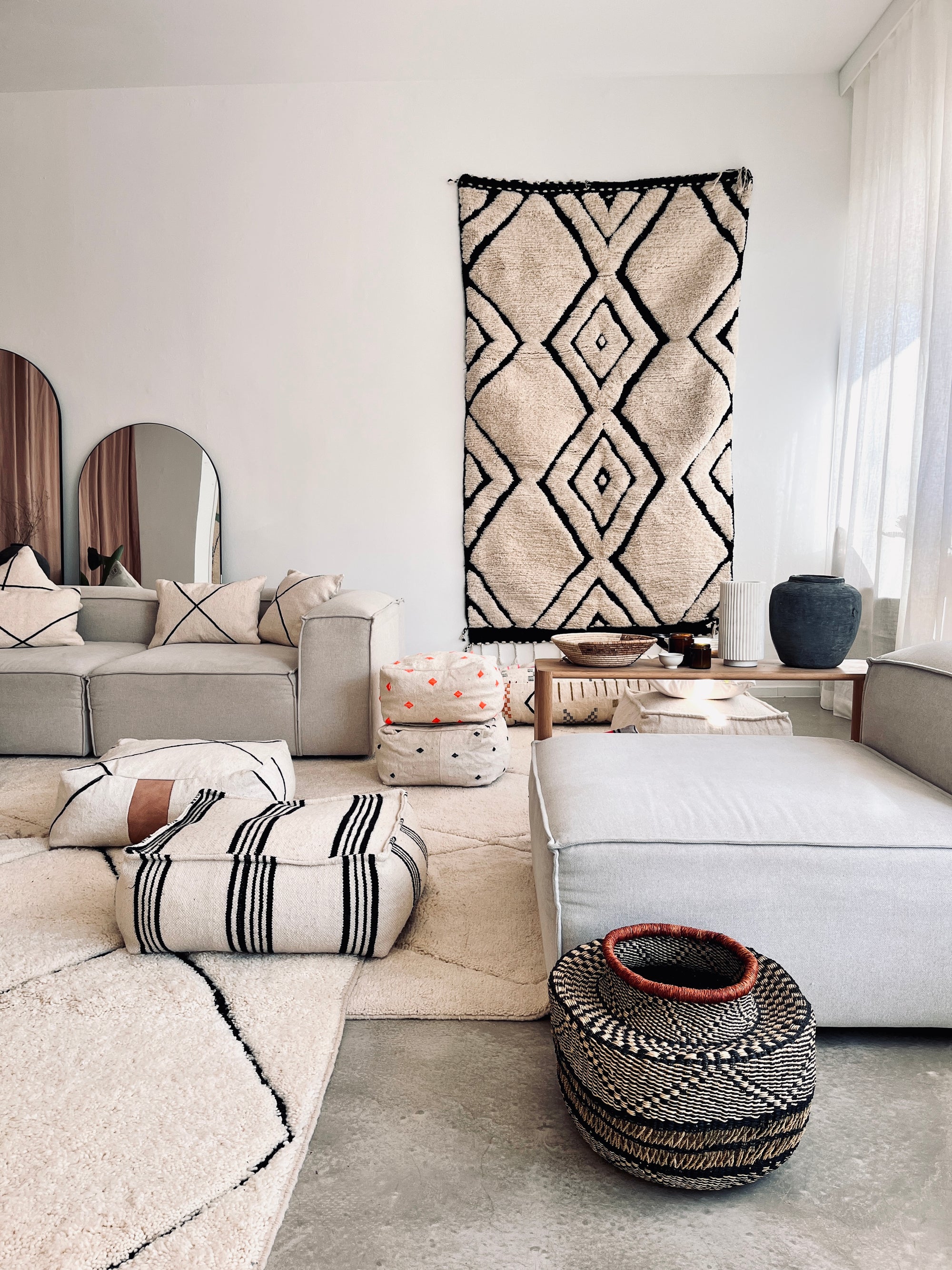 Ein modernes Wohnzimmer mit neutralen Tönen zeigt das LEY! TERMIN - SHOWROOM Sektionssofa, gemusterte Kissen, Bodenkissen, Poufs, geometrische Wandkunst, einen geflochtenen Korb, einen Couchtisch aus Holz und durchsichtige Vorhänge, die das Licht über einen weichen Teppich filtern.