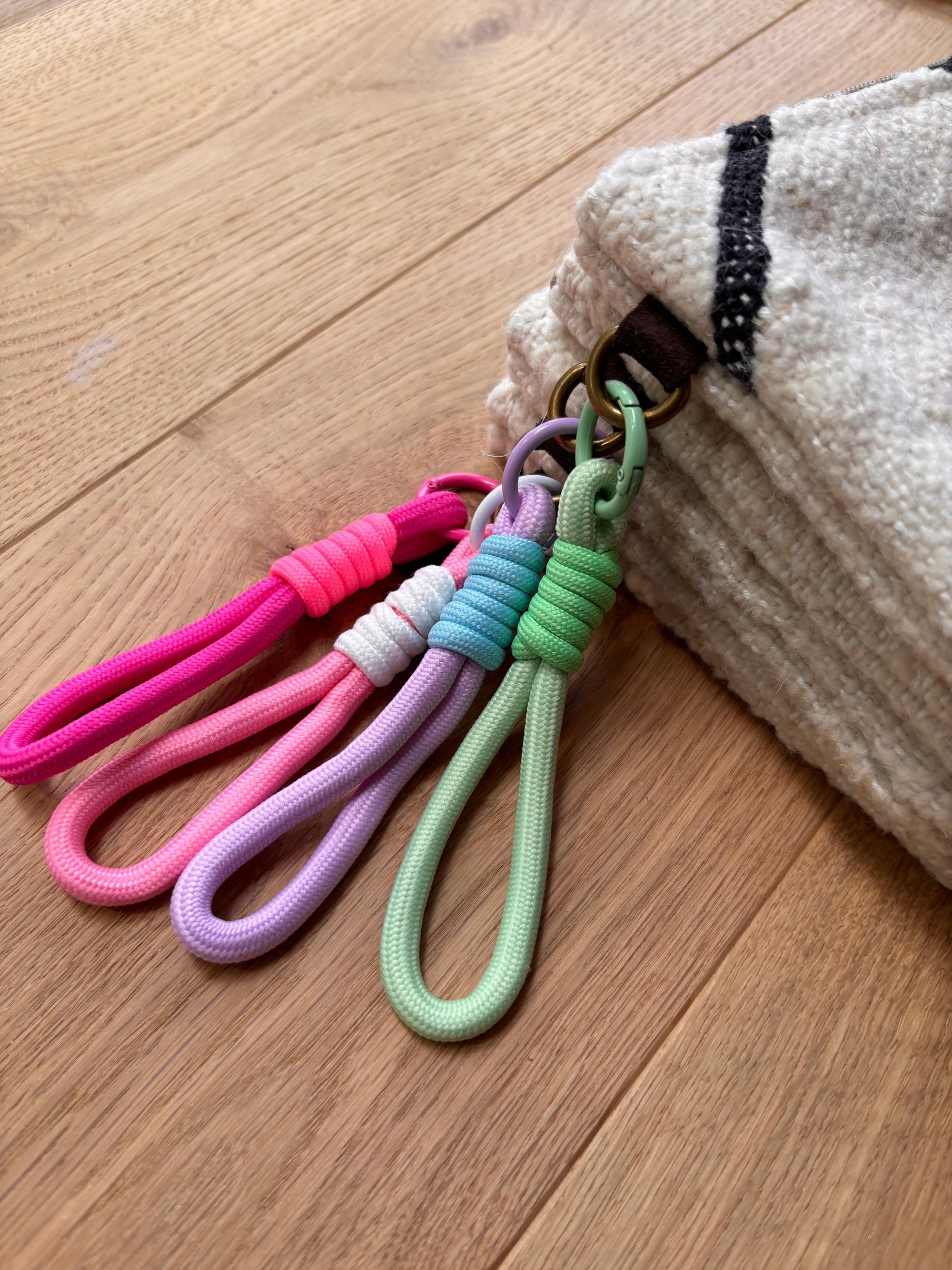 Vier bunte Schlüsselanhänger mit Schlaufen - rosa, lila, weiß und grün - hängen an einem Ring in der Ecke einer beigen LEY! Tasche - Classic Line - 15x20cm, hergestellt aus Schurwolle und auf einem Holzboden platziert.