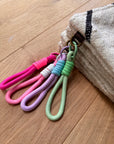 Vier bunte Schlüsselanhänger mit Schlaufen - rosa, lila, weiß und grün - hängen an einem Ring in der Ecke einer beigen LEY! Tasche - Classic Line - 15x20cm, hergestellt aus Schurwolle und auf einem Holzboden platziert.