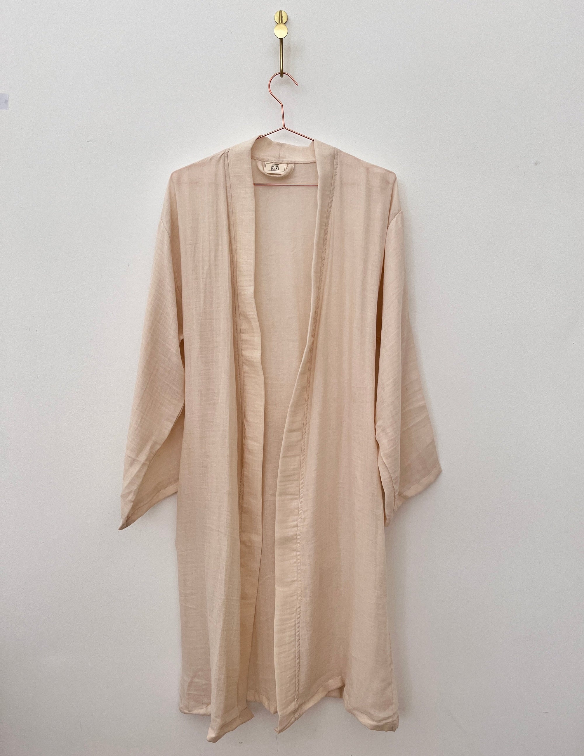Der KIMONO - nude von BIRLIK in Einheitsgröße aus weicher, beiger Baumwolle mit langen Ärmeln und knielangem Schnitt hängt an einem pinken Drahtbügel vor einer weißen Wand.