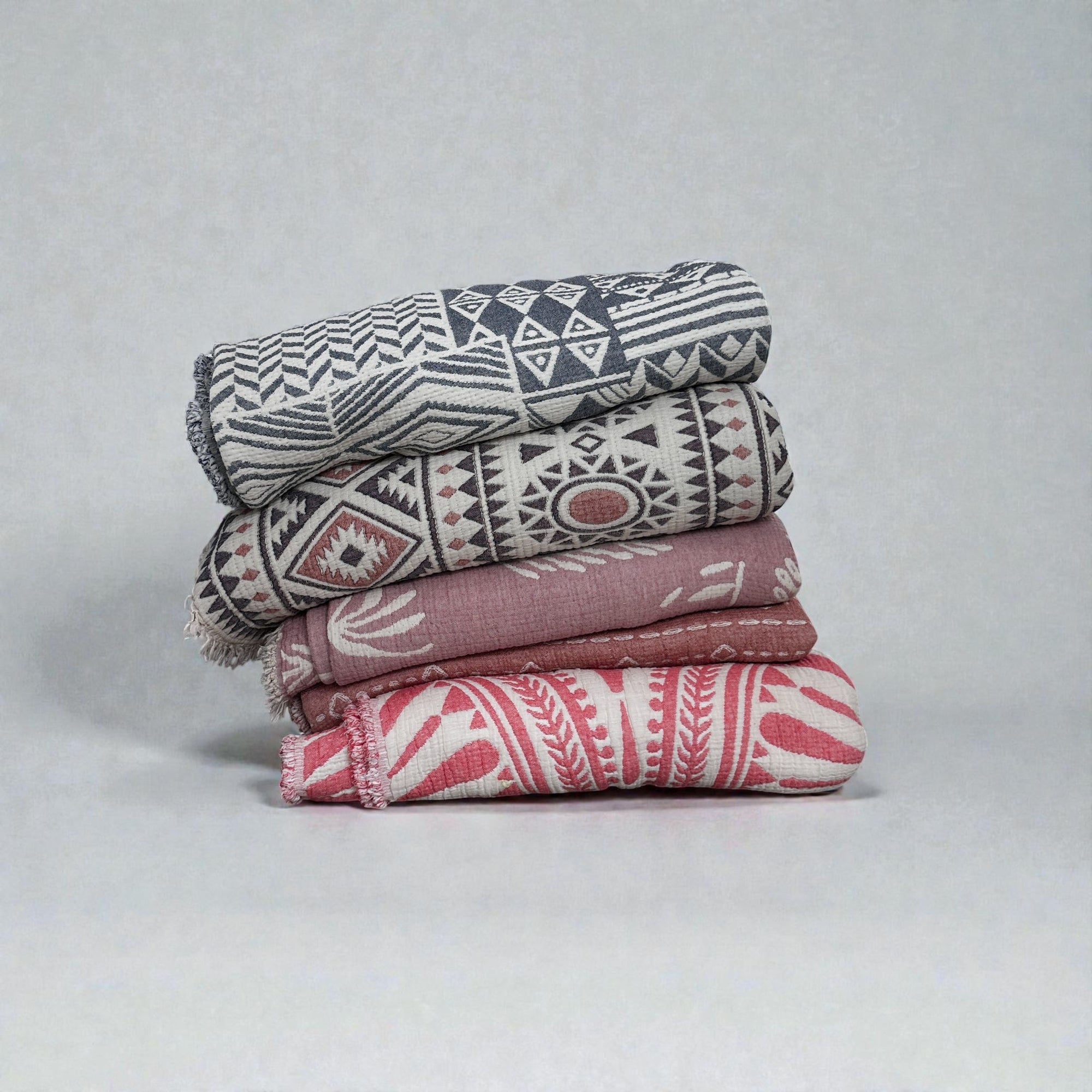 Vier gefaltete LEY! CALMA Musselin Decke (1,50 x 2,40 m) in Grau und Rosa, mit luftigem, atmungsaktivem Stoff und geometrischen oder botanischen Boho-Designs, werden vor einem einfarbigen Hintergrund gestapelt.
