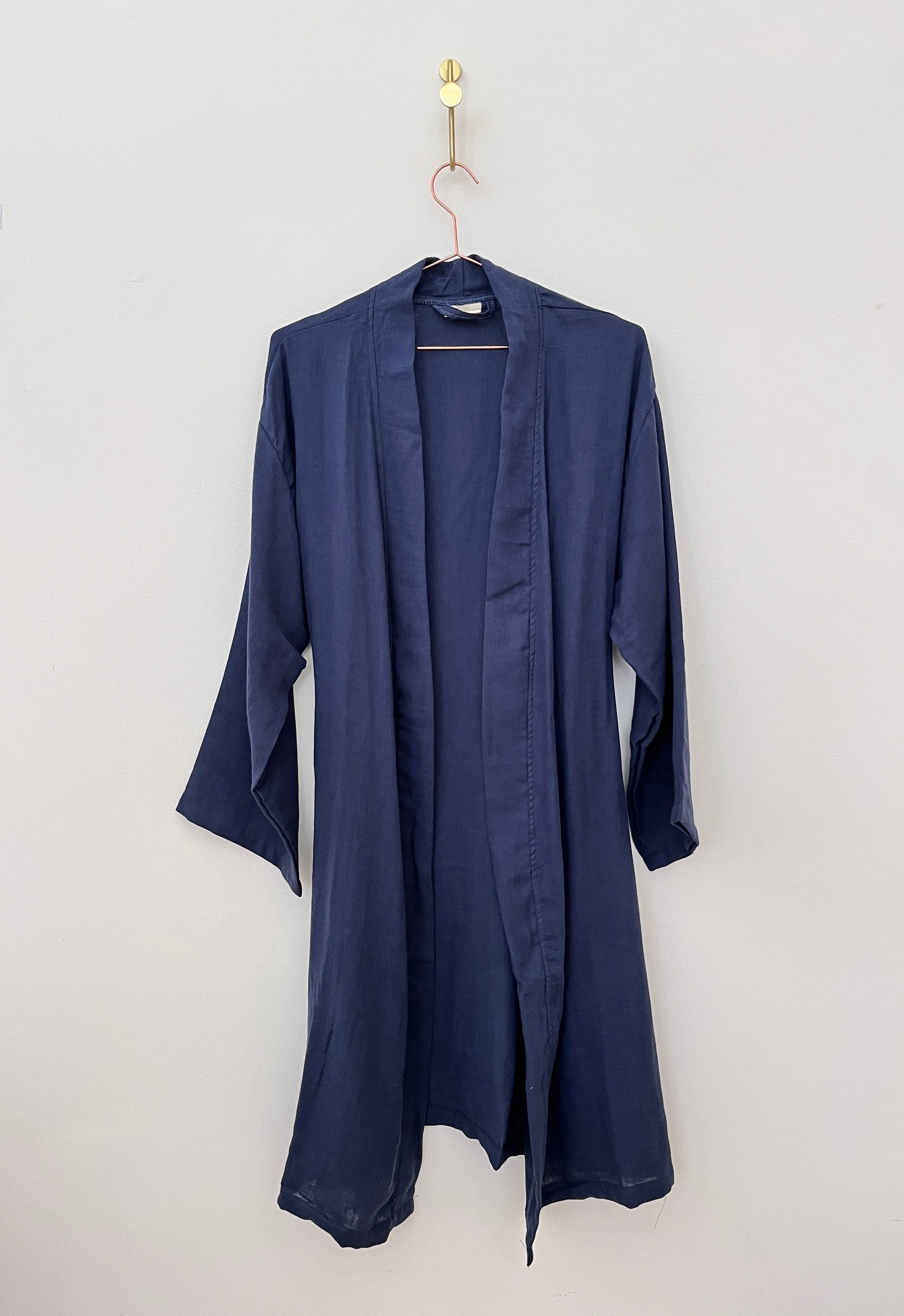 Der KIMONO - Dunkelblau von BIRLIK ist ein marineblauer, knielanger Bademantel aus Baumwolle mit langen Ärmeln und einer offenen Vorderseite. Er hat eine lockere Passform, ist in Einheitsgröße erhältlich und wird an einem goldenen Haken vor einer schlichten weißen Wand aufgehängt.