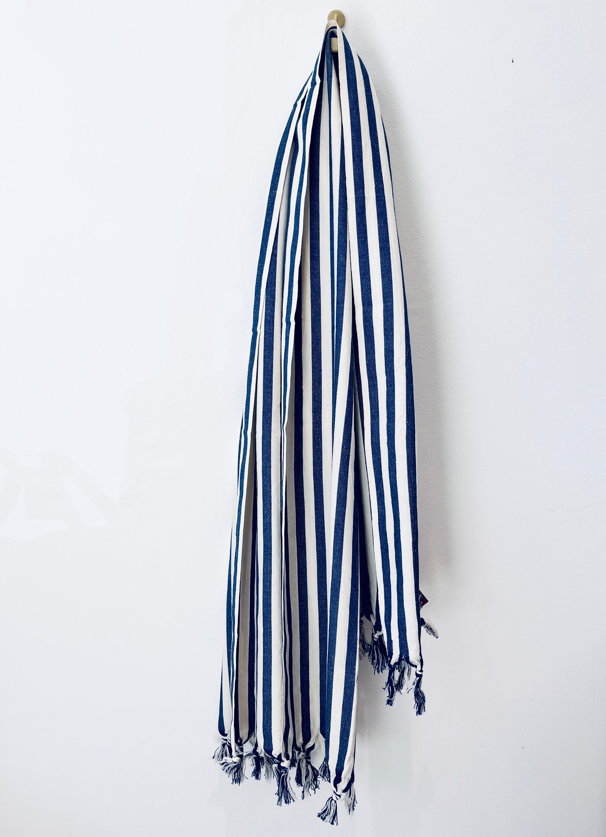 Ein blau-weiß gestreiftes Plaid - 90x180 blau von BIRLIK mit ausgefransten Enden hängt an einem einzelnen Haken an einer weißen Wand.