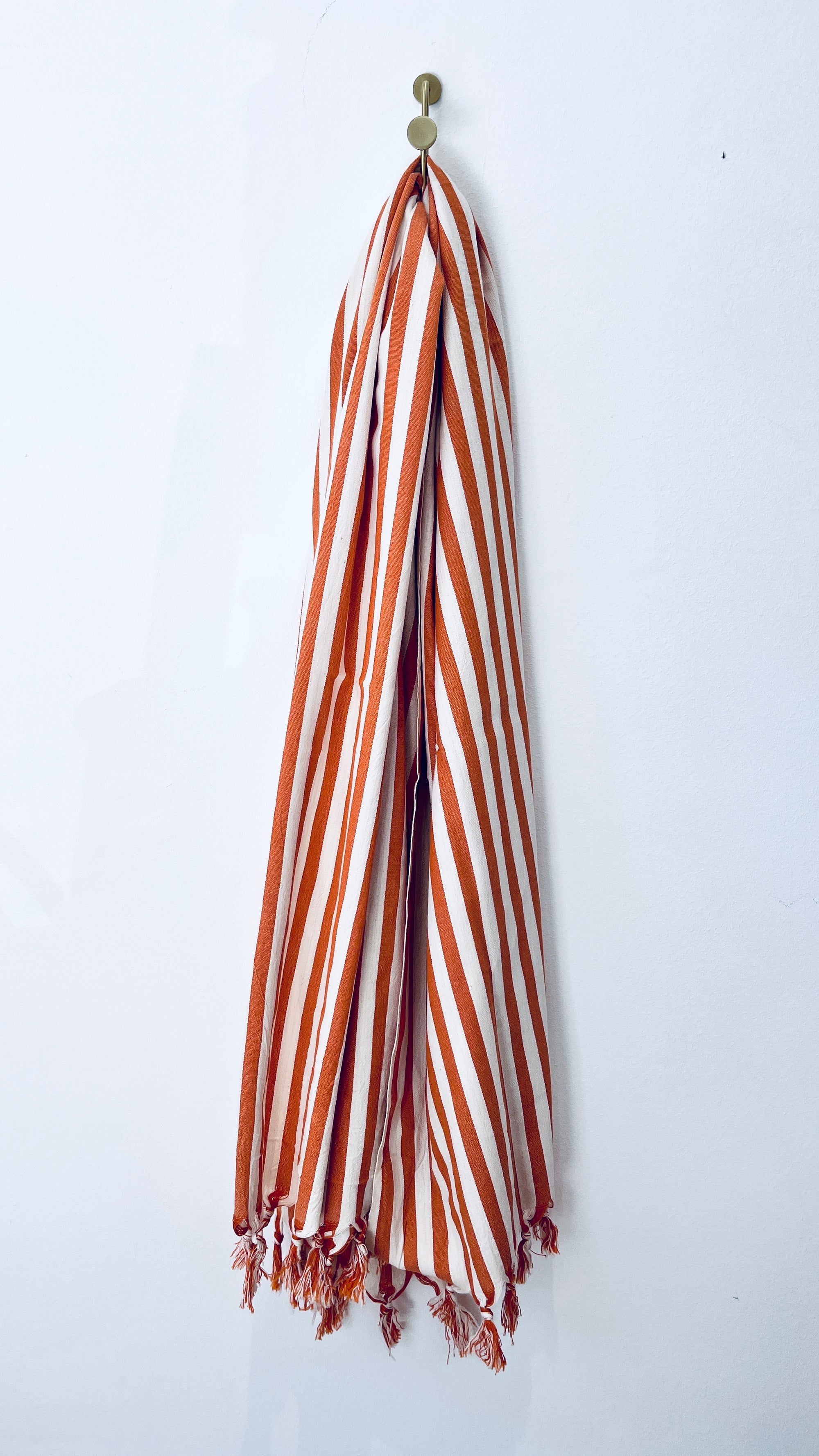 A Plaid - 90x180 terracotta von BIRLIK hängt an einem goldenen Haken an einer schlichten weißen Wand. Die roten und weißen Streifen mit Fransen an den Enden bilden einen auffälligen Anblick.