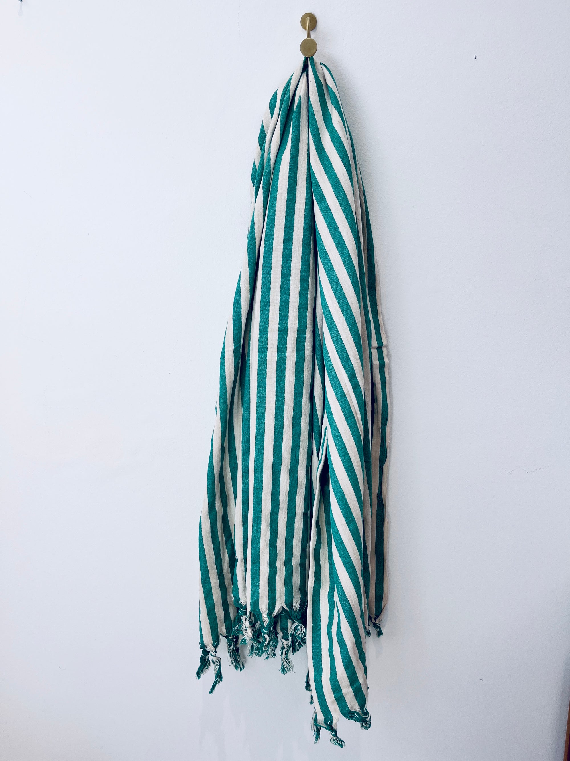 Das grün-weiß gestreifte Plaid BIRLIK - 90x180 grün mit ausgefransten Enden hängt an einem einzigen runden Haken an einer schlichten weißen Wand.