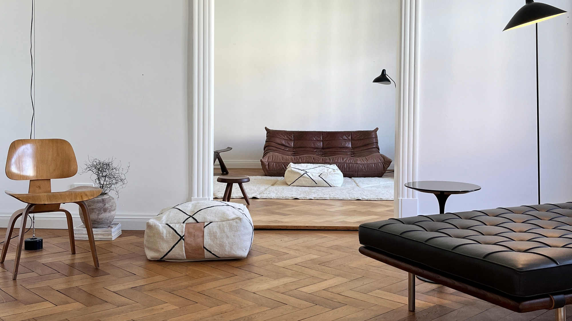 Ein modernes Wohnzimmer mit Fischgräten-Holzboden, einer Ledercouch, einem Holzstuhl, einem Hocker und einem niedrigen braunen Sofa. Ein großer Spiegel reflektiert den Raum und unterstreicht sein minimalistisches Dekor.