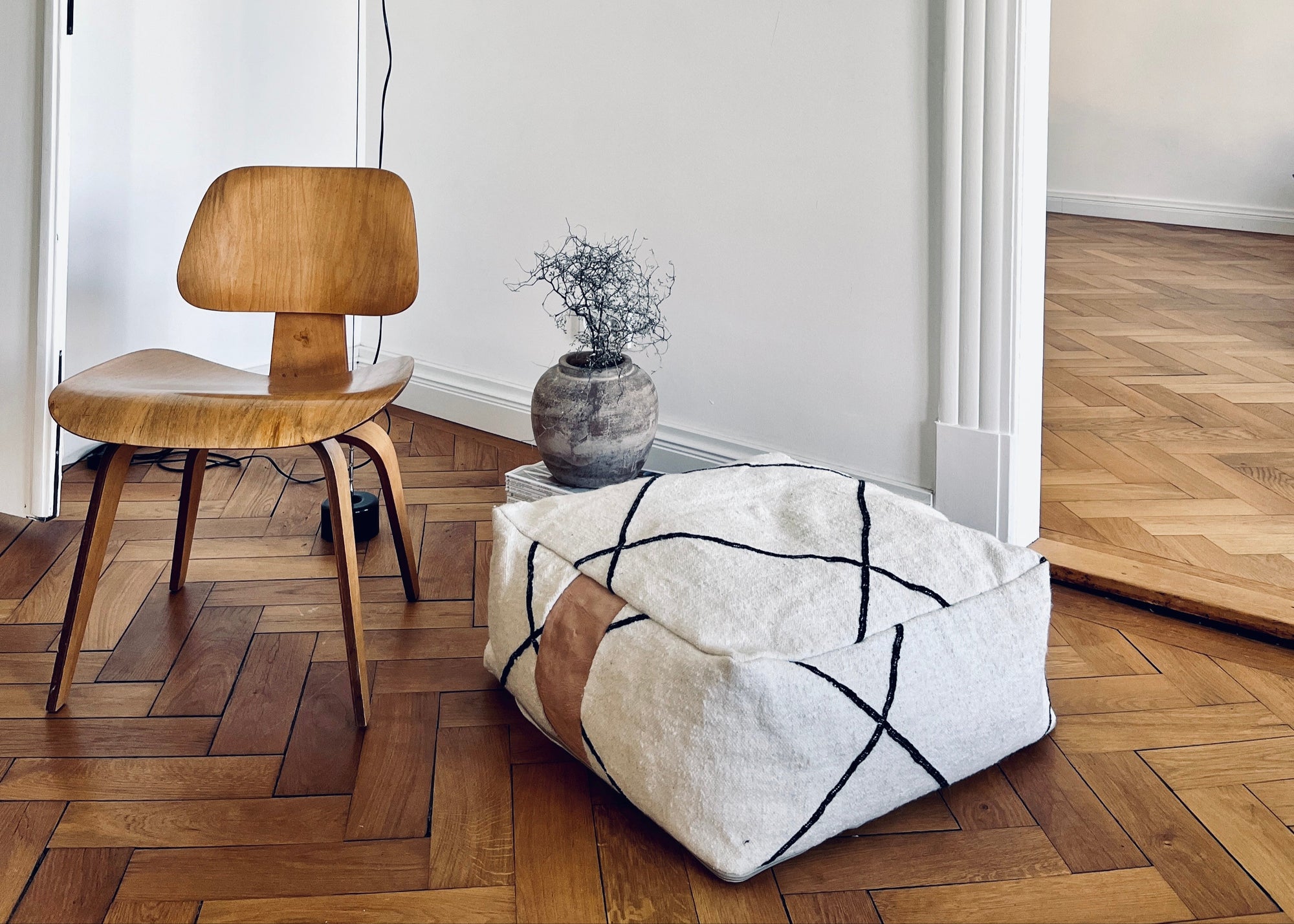 Ein minimalistischer Raum mit einem Holzstuhl, einem großen Kissen mit weißem Bezug und schwarzen Linien, einer Keramikvase mit getrockneten Zweigen und einem Holzboden mit Fischgrätenmuster.