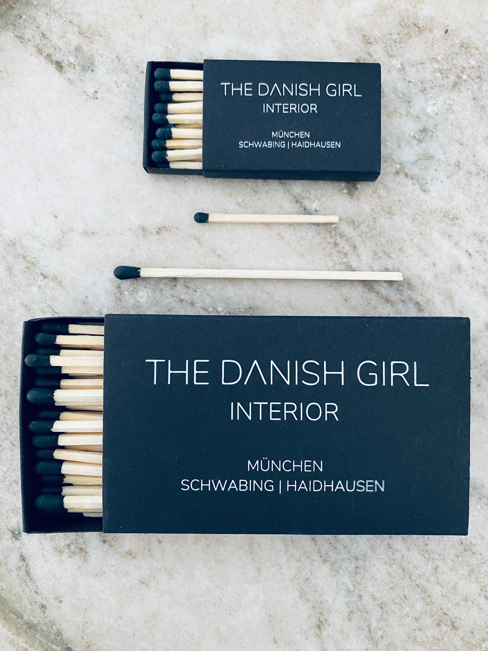 Zwei schwarze Streichhölzer-Schachteln von LEY!, eine große und eine kleine, sind mit "The Danish Girl Interior München Schwabing | Haidhausen"-Etiketten und Holzstreichhölzern versehen. Beide Schachteln sind individuell gestaltbar und ein gebrandetes Accessoire.