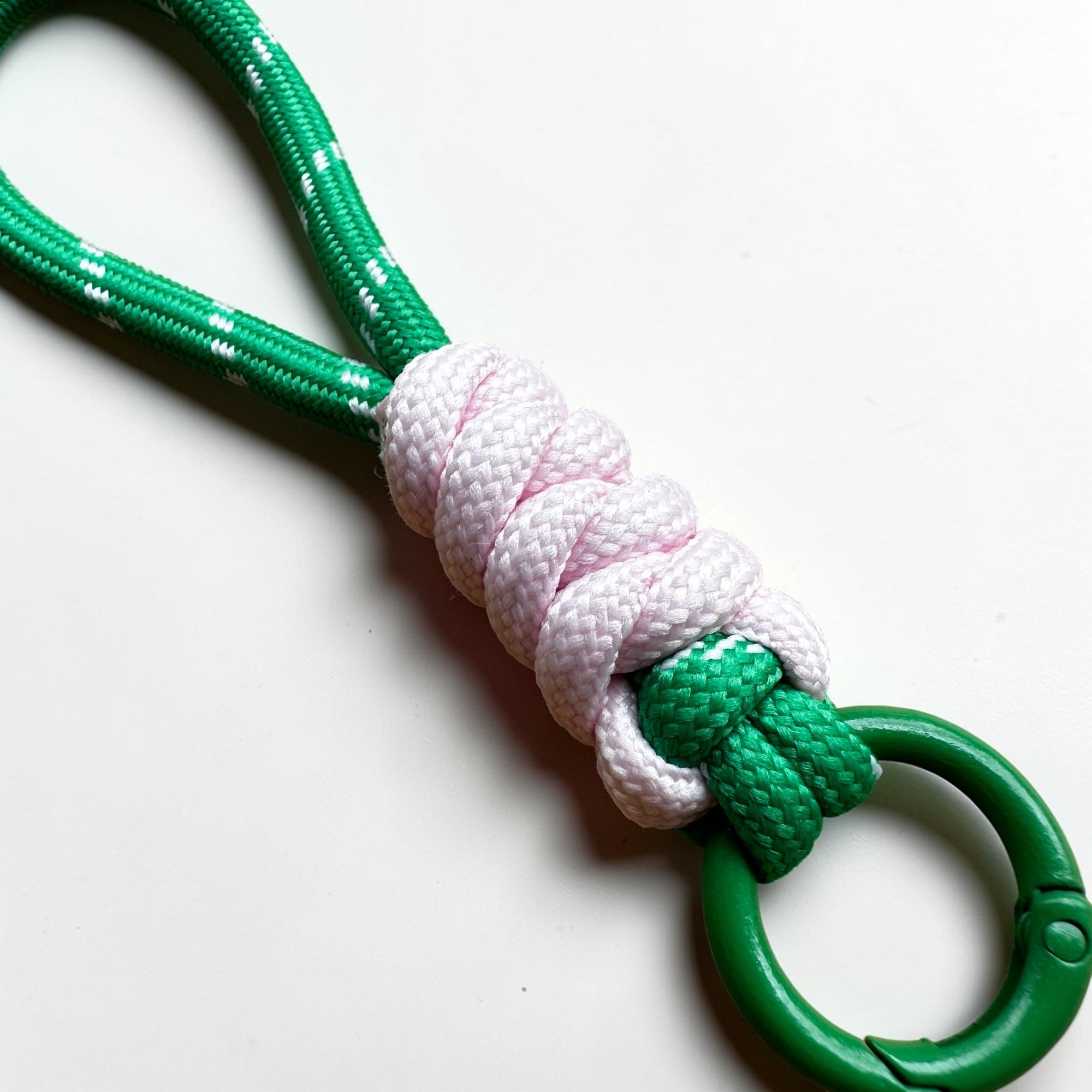Eine Nahaufnahme des LEY! Schlüsselanhänger - grün - weiß - rosa, mit geschlungenem und geknotetem grün-weißem Seil mit einem rosa-weißen Knoten in der Mitte, einem grünen Ring und ideal für Taschen, auf einer weißen Fläche.