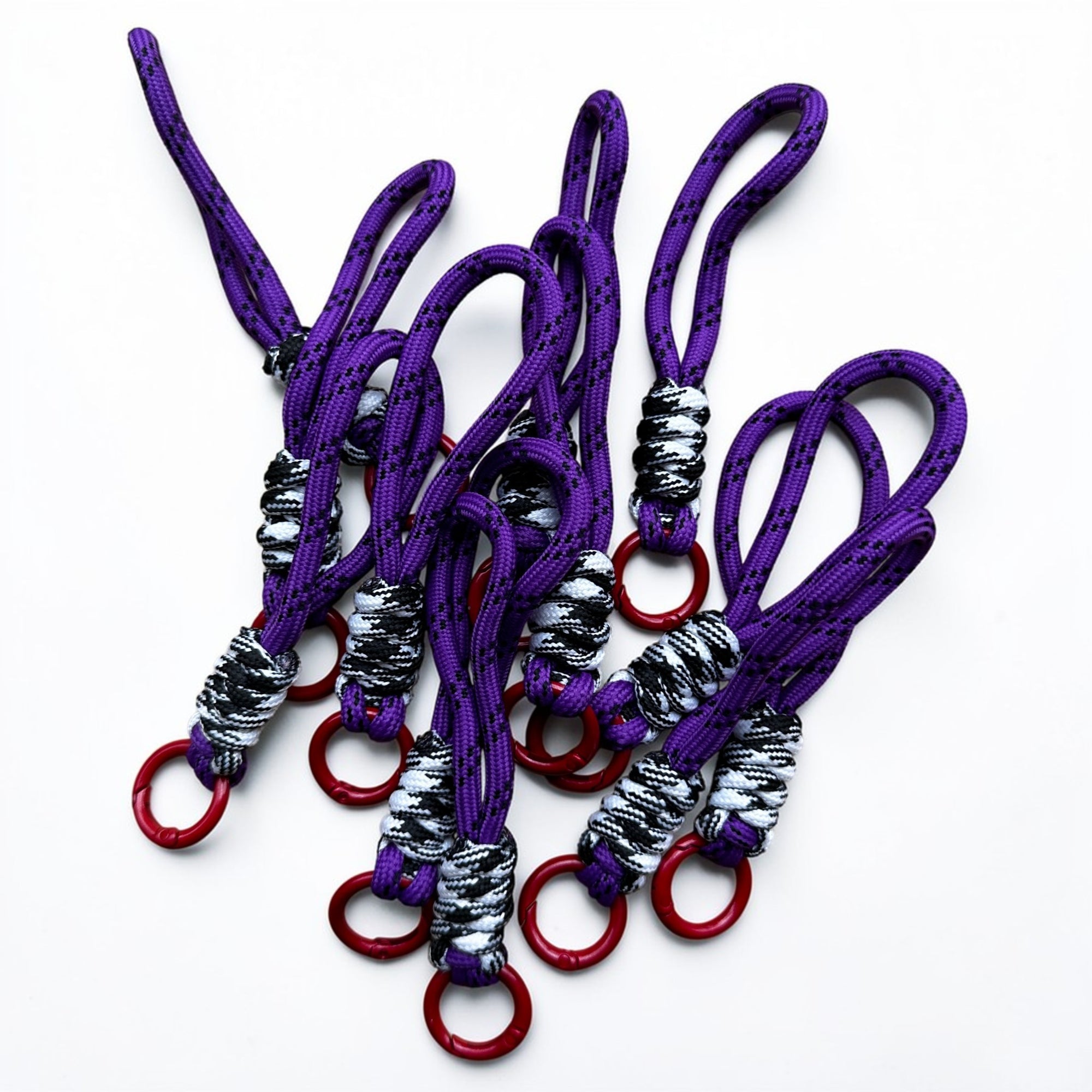 Der Schlüsselanhänger - lila - schwarz - weiß - rot von LEY! hat lilafarbene Paracord-Schlaufen mit schwarzen Mustern, die mit schwarz-weißer Kordel umwickelt und mit einem roten Metallring versehen sind - ein auffälliges Accessoire für jede Tasche.