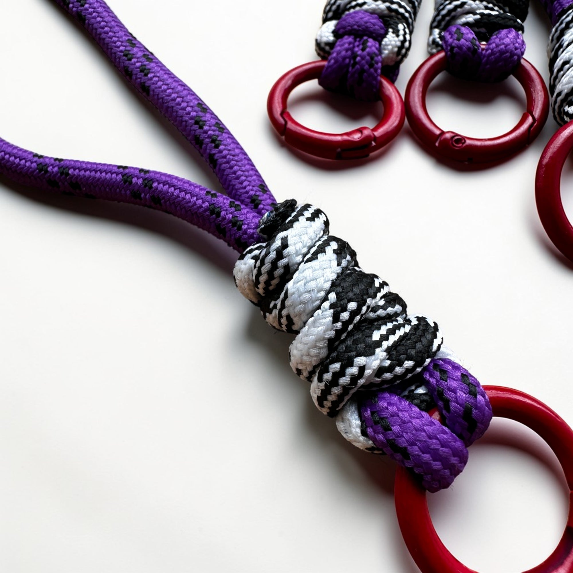 Nahaufnahme des LEY! Schlüsselanhänger - lila, schwarz, weiß &amp; rot - mit geknoteten Seilen, die durch rote Ringe geschlungen sind, ideal als kreatives Accessoire für Ihre Tasche.