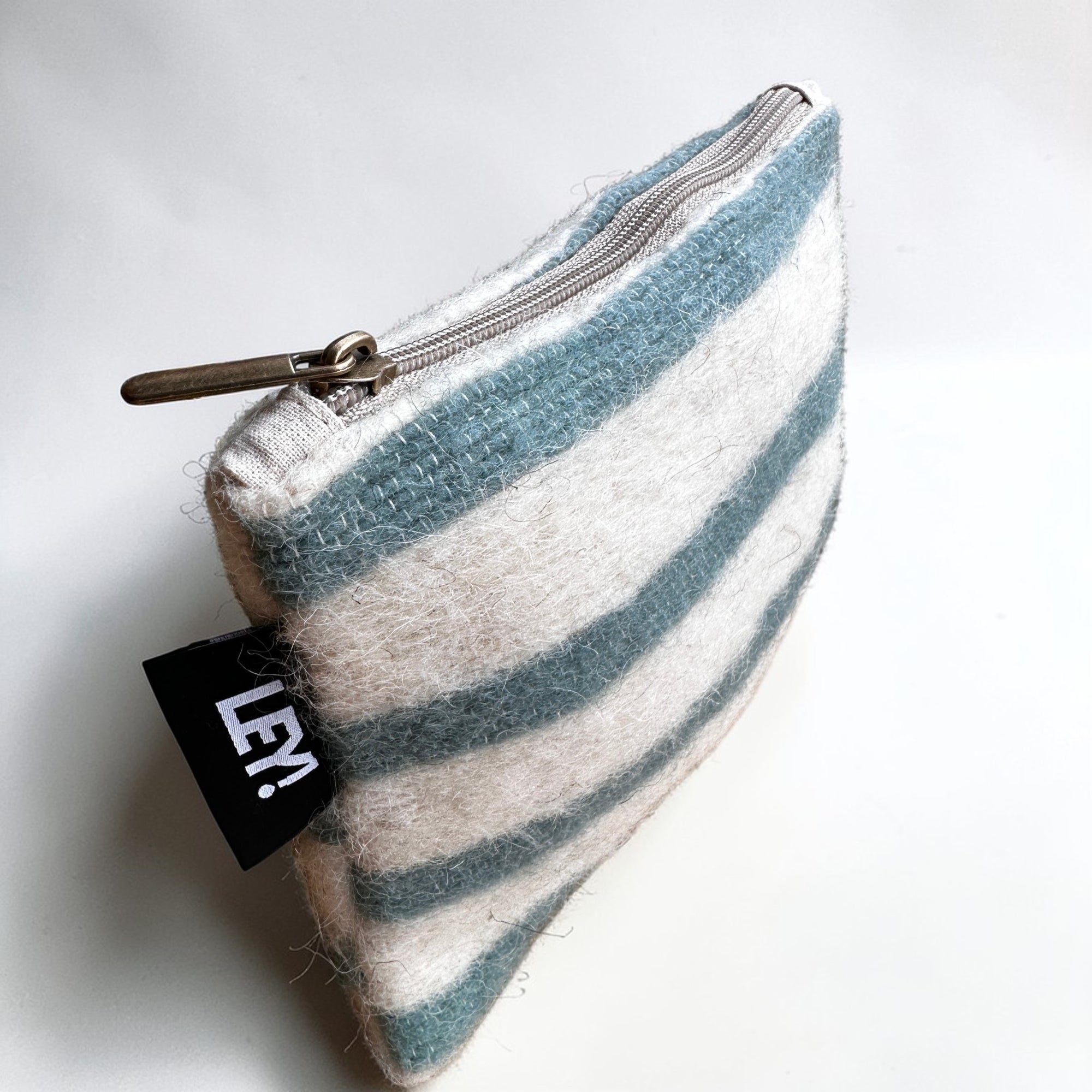 Die LEY! Tasche - WINTER EDITION - LAGUNE (15 x 20 cm) ist eine kleine Reißverschlusstasche aus strukturierter Schurwolle mit blauen und weißen Streifen und einem schwarzen "LEY!"-Anhänger - perfekt für den Winter und inspiriert von Handarbeit Marrakesch.