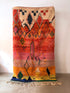 Der "Berber Teppich - Nina - 1,50 x 2,50" von LEY! ist ein farbenfroher handgefertigter Teppich aus weicher Schurwolle mit abstrakten geometrischen Mustern und Fransen, der senkrecht an einer weißen Wand hängt.