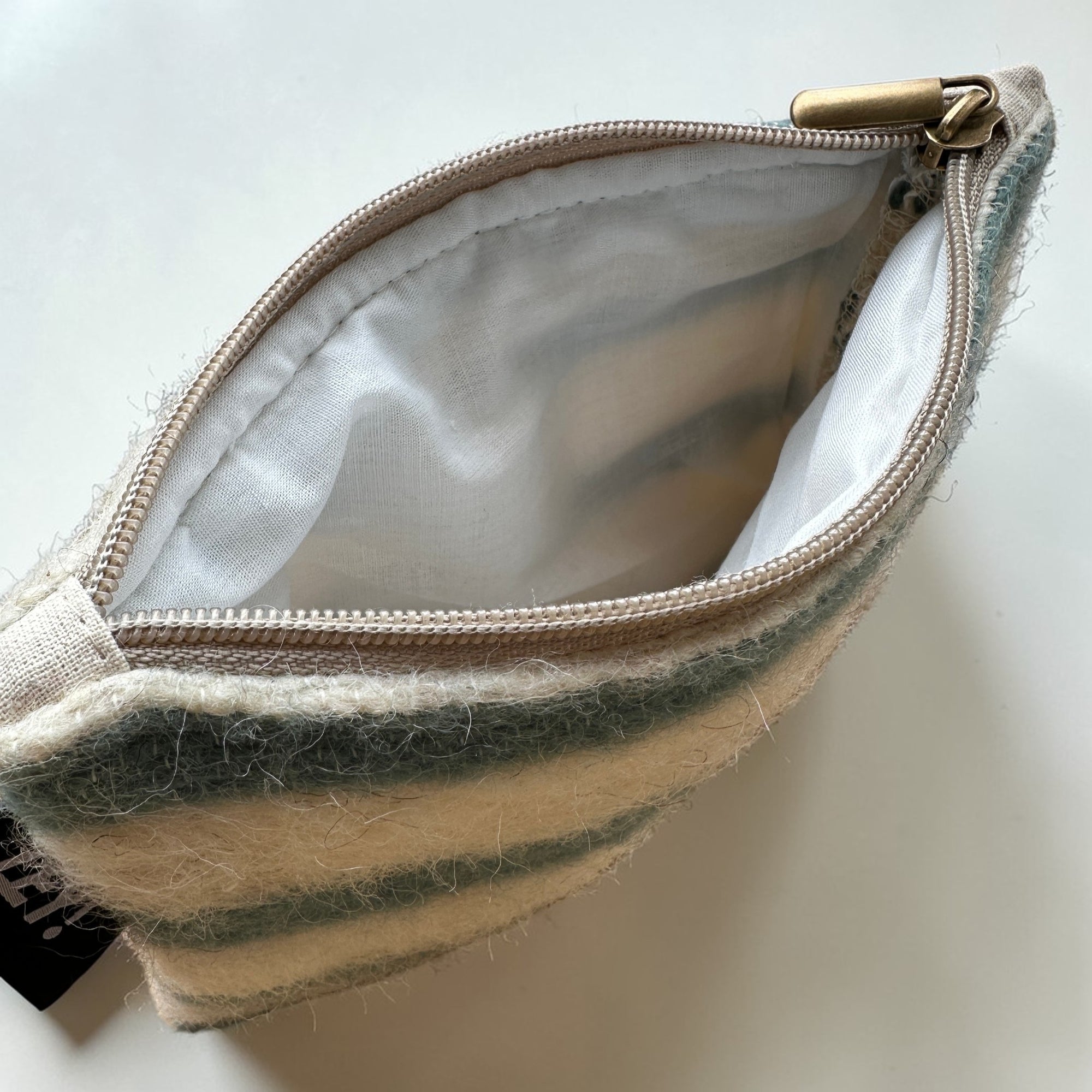 Die LEY! Tasche - WINTER EDITION - LAGUNE (15 x 20 cm) ist eine kleine, in Marrakesch gefertigte Reißverschlusstasche aus weicher, gestreifter Schurwolle mit weißem Innenfutter und Metallreißverschluss, die geöffnet auf einer weißen Fläche abgebildet ist.