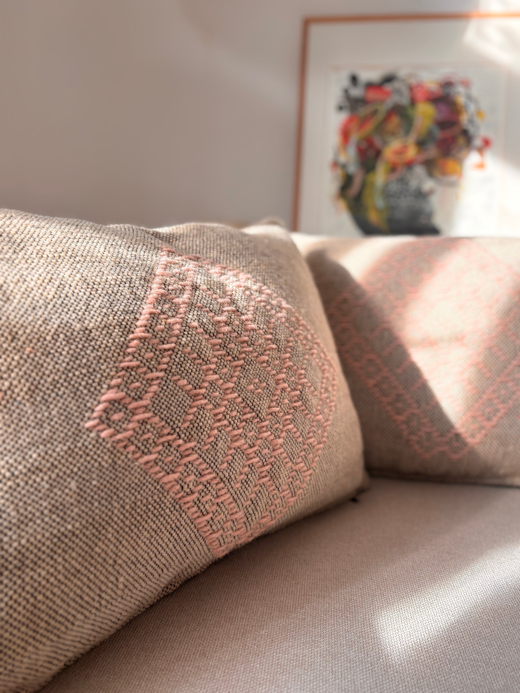Eine beigefarbene Couch mit dem Heritage Line Kissen in Rosé (40 x 60 cm) von LEY! mit zartrosa Stickerei wird in Sonnenlicht getaucht, wobei im Hintergrund ein lebhaftes gerahmtes Kunstwerk teilweise sichtbar ist.