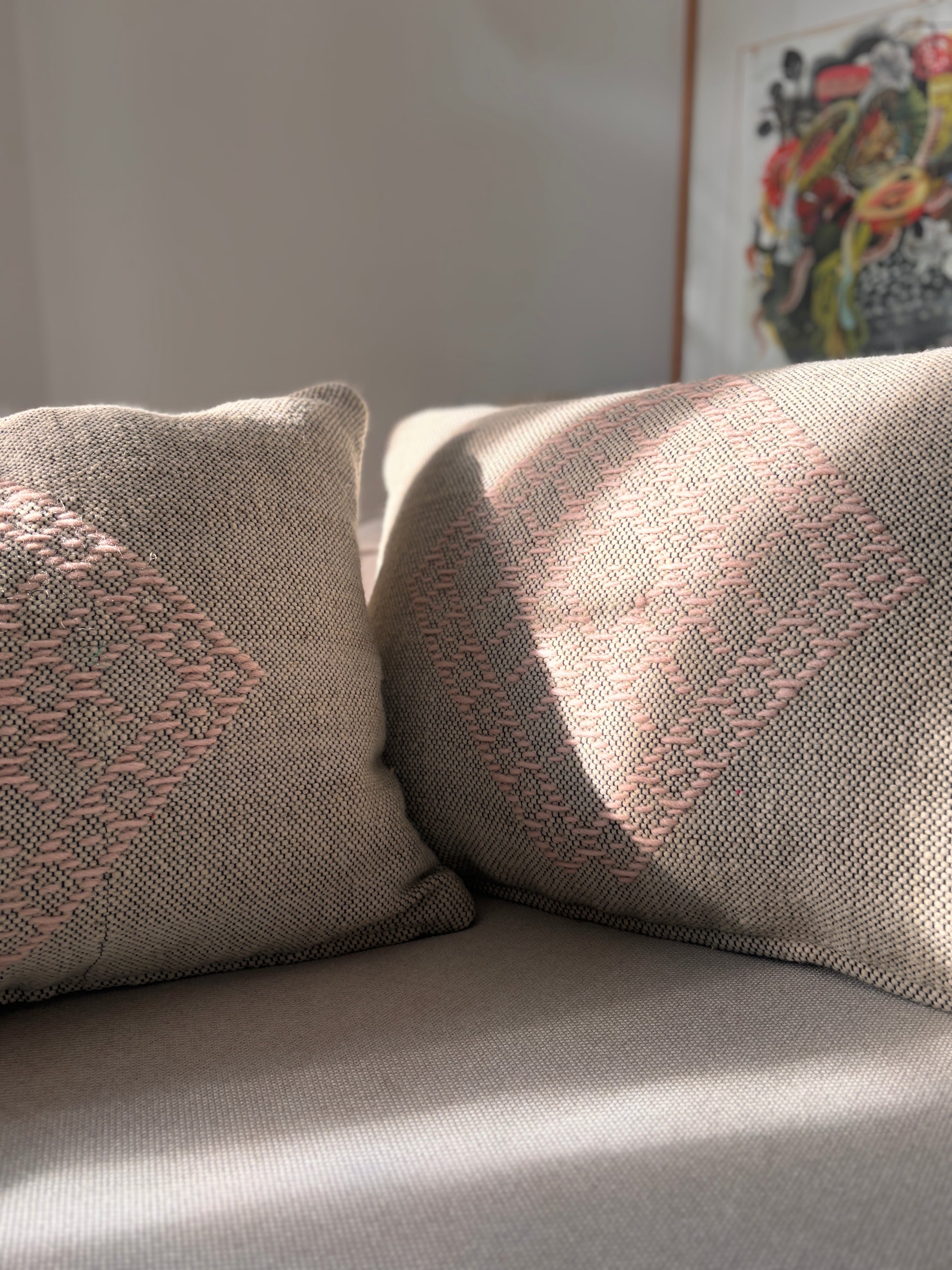 Zwei Heritage Line Kissen - Rosé (40 x 60 cm) von LEY! mit pinkem Diamantmuster liegen auf einem hellen Sofa im Sonnenlicht, während im Hintergrund ein abstraktes Kunstwerk zu sehen ist.