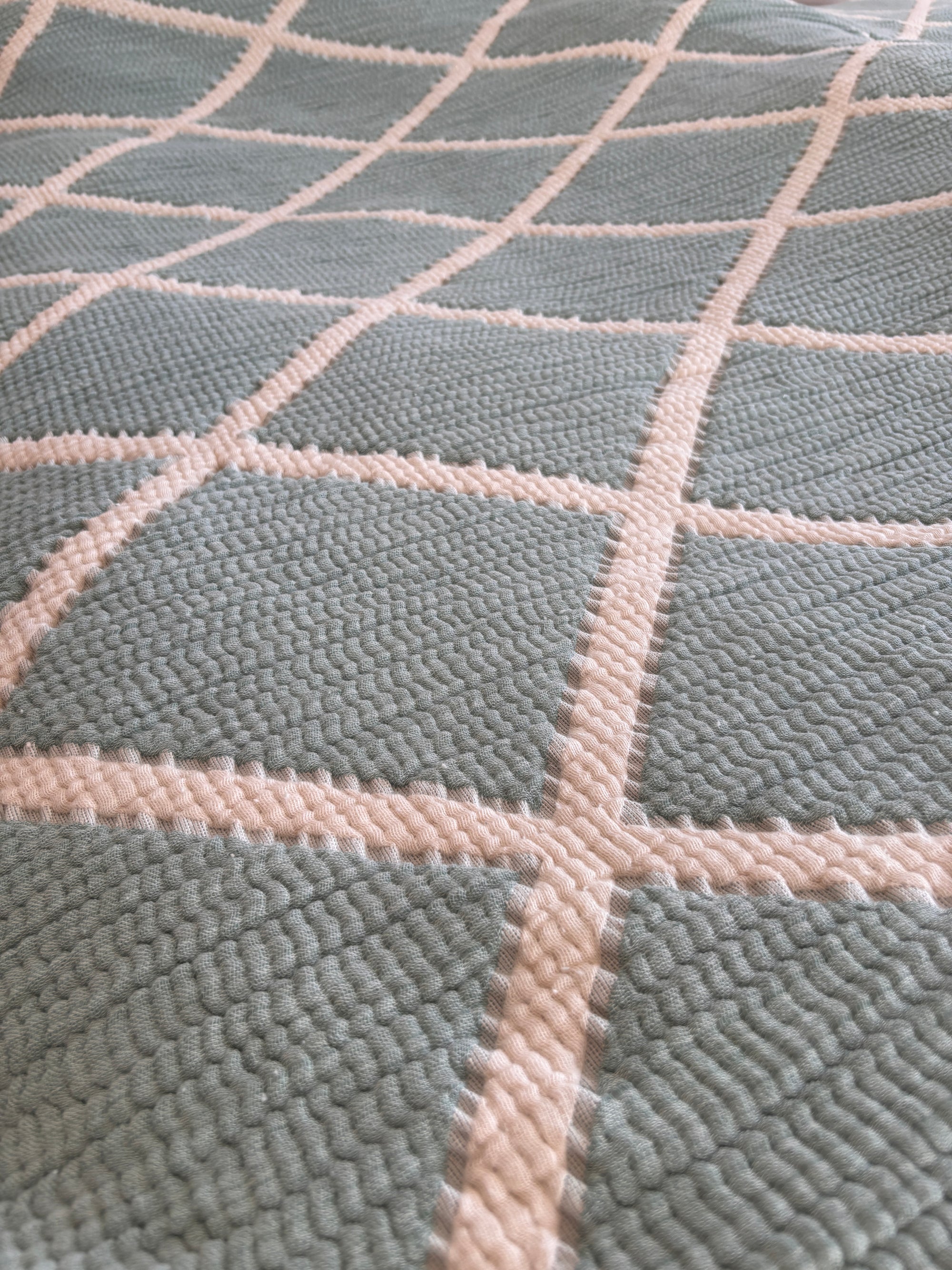 Eine Nahaufnahme der LEY! Musselin Decke - Salbei-Weiß (2,20 x 2,50 m) zeigt ihre gesteppte Oberfläche und das erhabene cremefarbene Gitter vor einem zartblauen Hintergrund, der dem Design einen subtilen geometrischen und Boho-Touch verleiht.