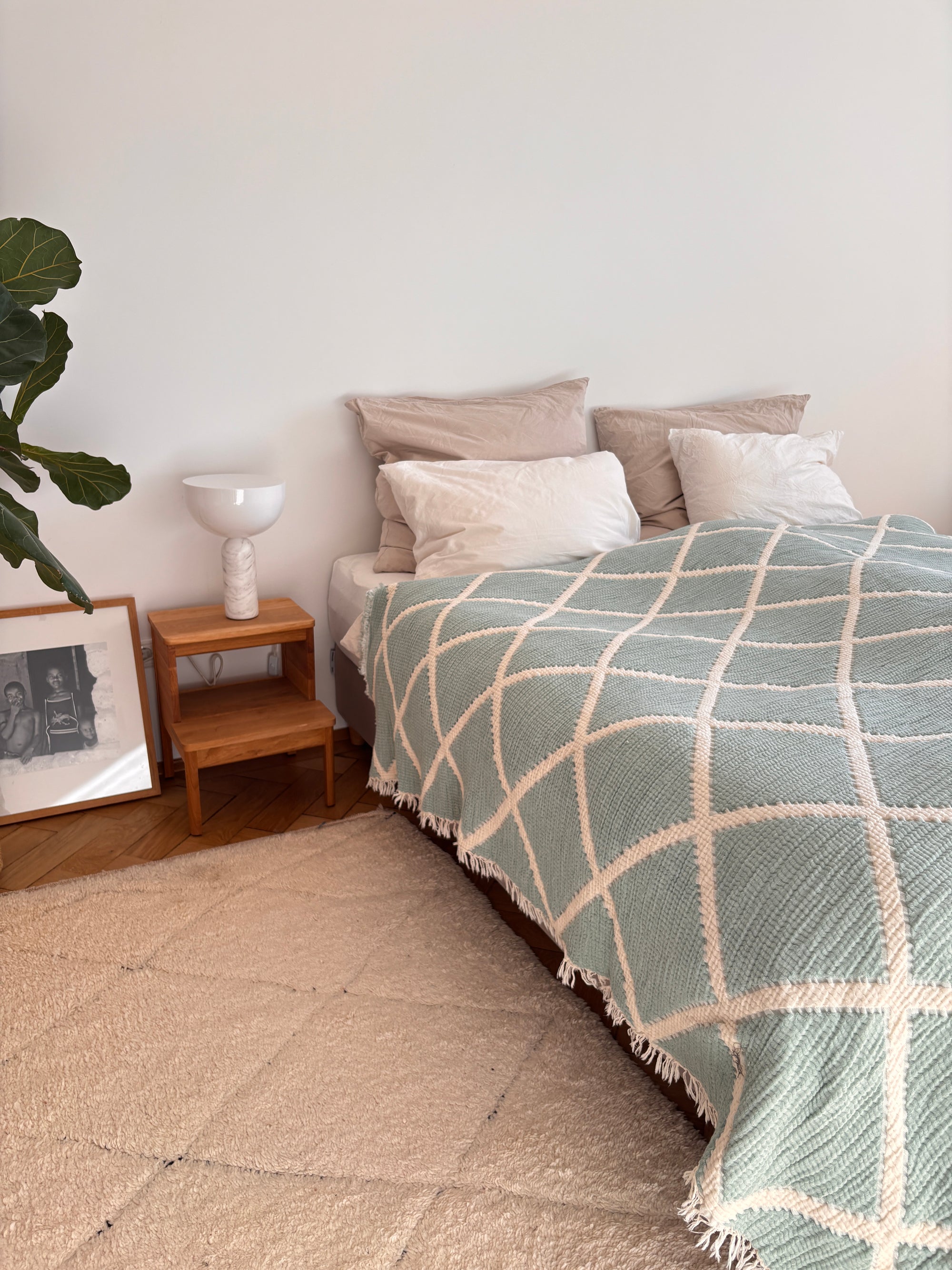 Ein ordentlich gemachtes Bett mit beigen und weißen Kissen wird mit der LEY! Musselin Decke in Grau-Weiß (2,20 x 2,50 m), die für Boho-Flair sorgt. Auf einem hölzernen Nachttisch steht eine weiße Lampe, daneben stehen eine Blattpflanze und ein gerahmtes Schwarz-Weiß-Foto.