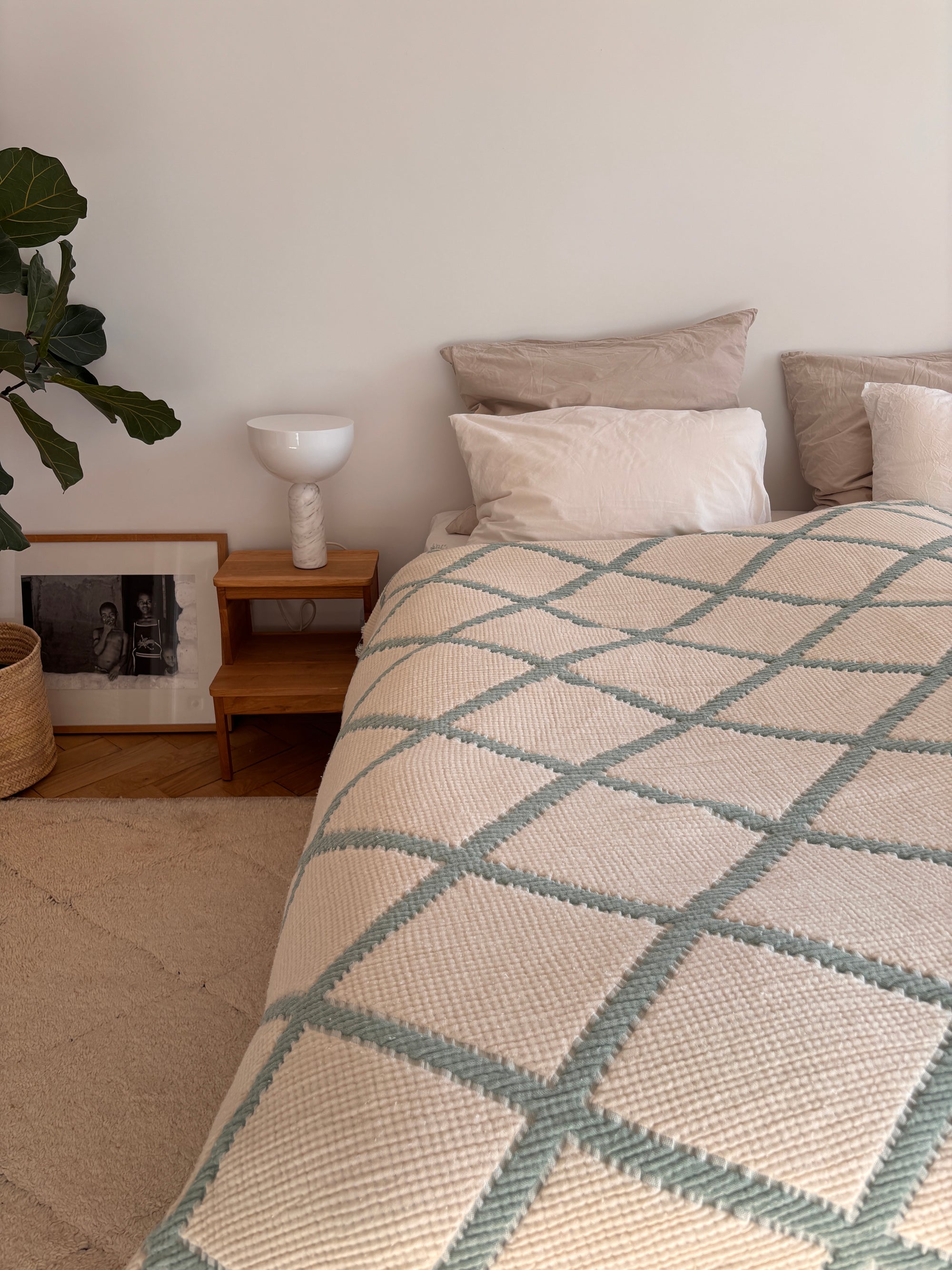 Ein gemütliches Schlafzimmer mit einem hübsch gemachten Bett, das mit der LEY! Musselin Decke in Salbei-Weiß (2,20 x 2,50 m), neutralen Kissen, einem hölzernen Nachttisch mit einer weißen Lampe, einer Topfpflanze und einem gerahmten Foto auf dem Boden.