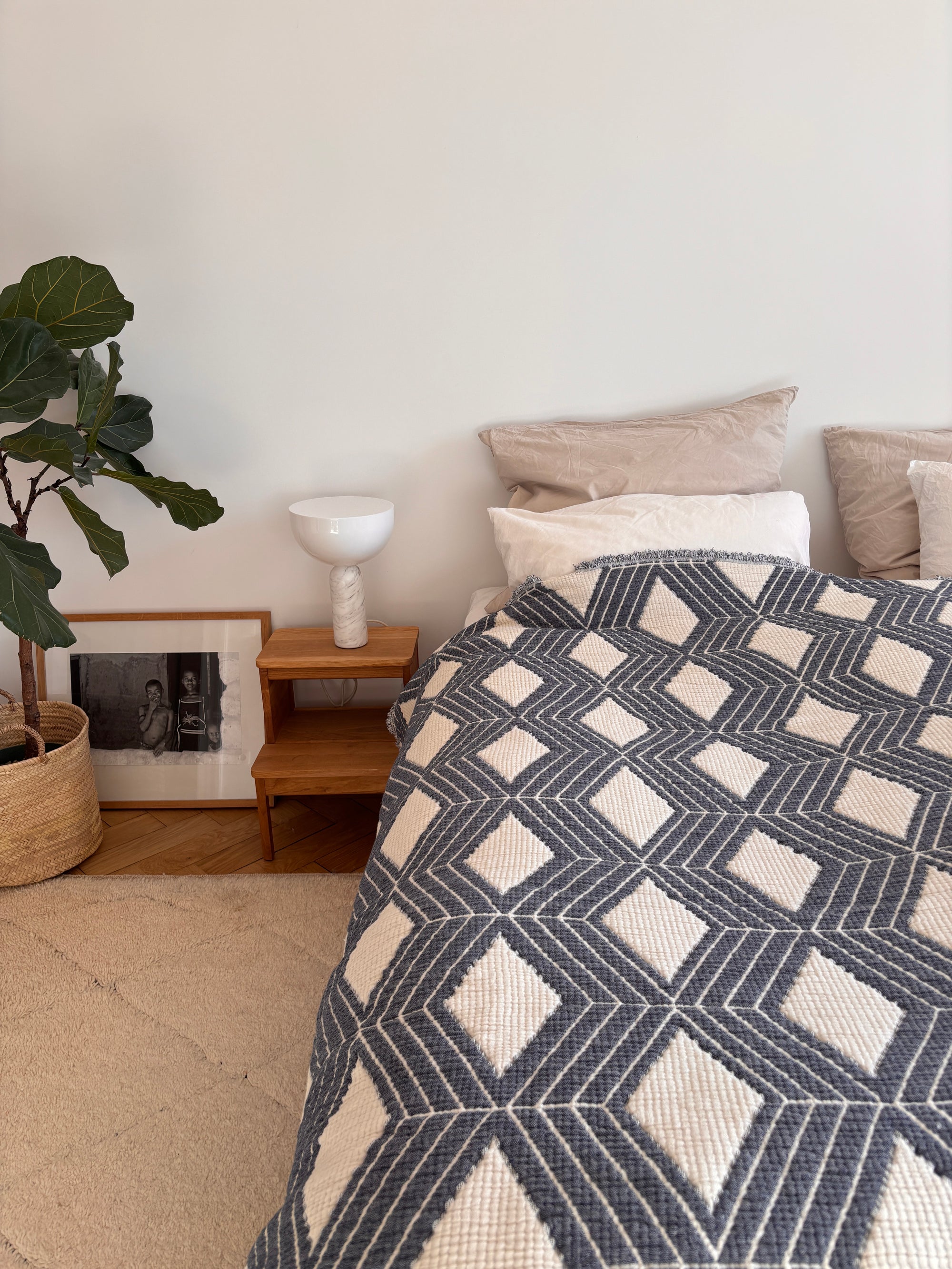 Ein modernes Schlafzimmer mit einem Hauch von Boho-Design, mit einem Bett, das mit der LEY! Musselin Decke - Blau-Weiß (2,20 x 2,50 m), beigen Kissen, einem hölzernen Nachttisch mit einer weißen Lampe, einer Topfpflanze und einem gerahmten Foto an der Wand.