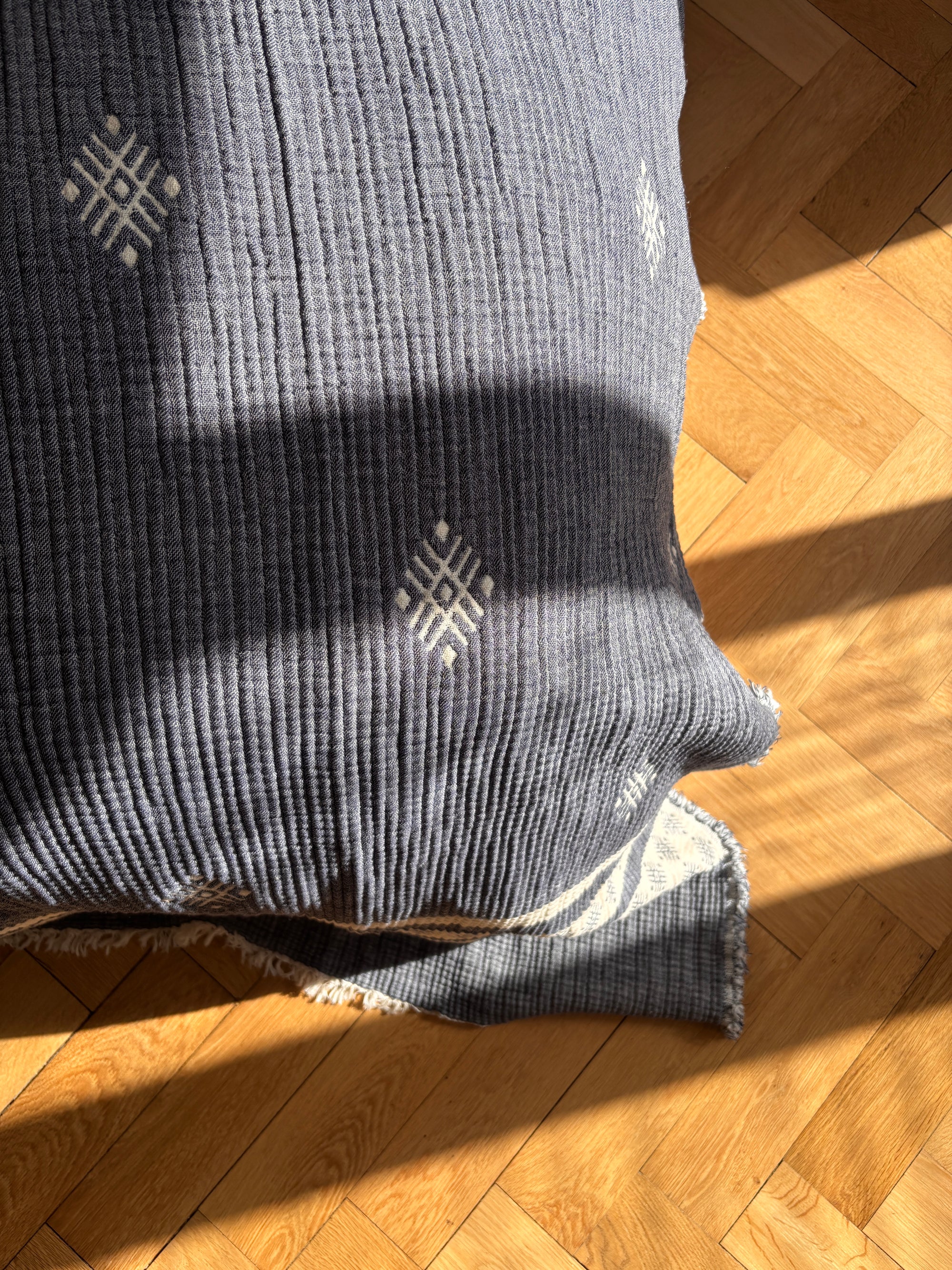 Die LEY! Musselin Decke - Blau-Weiß (2,20 x 2,50 m) liegt teilweise gefaltet auf einem hellen Parkettboden im Sonnenlicht und setzt einen gemütlichen, stilvollen Akzent in jeder Boho-Einrichtung.