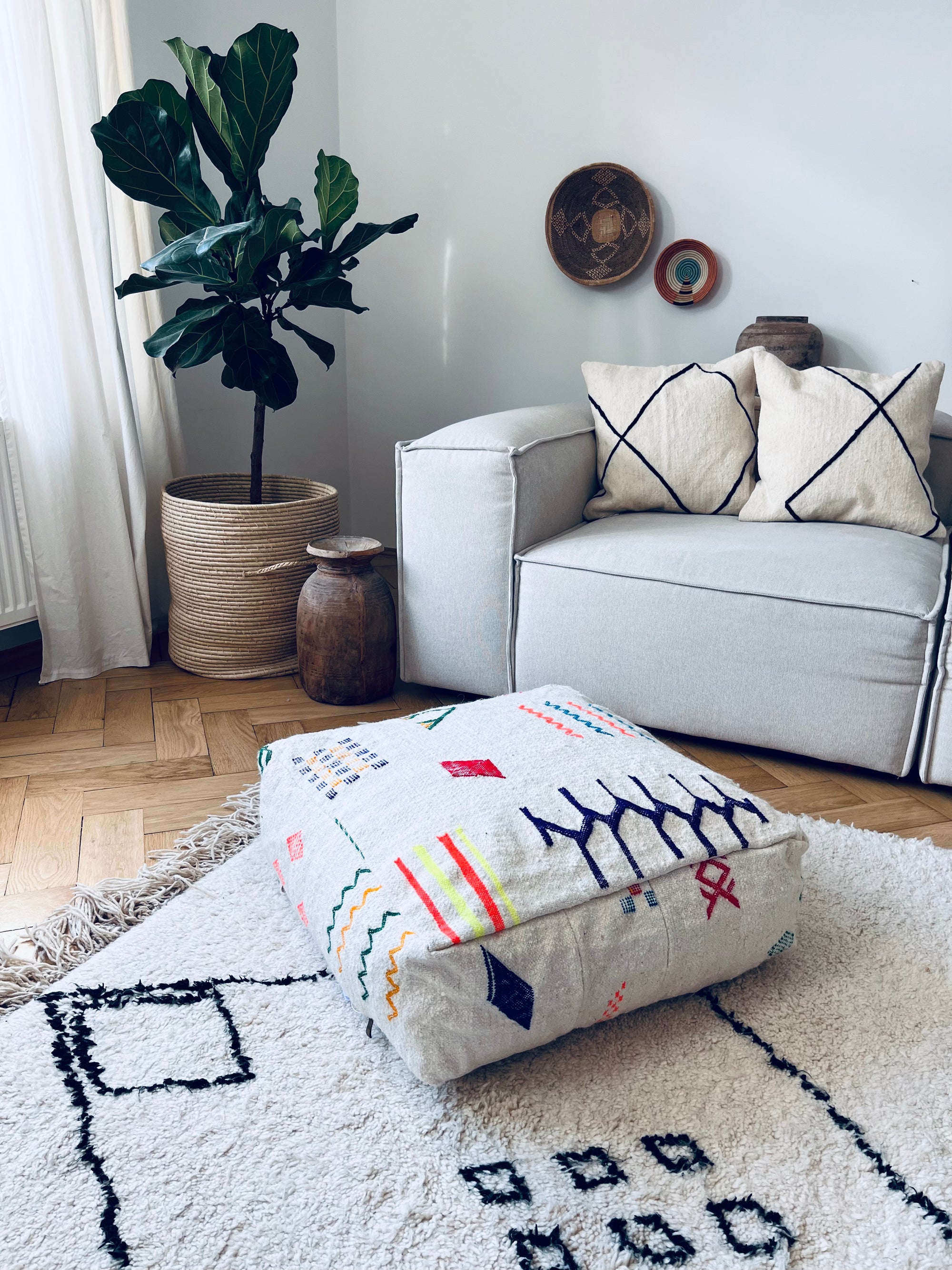 Ein modernes Wohnzimmer mit einer großen Topfpflanze, einem hellgrauen Sofa mit zwei gemusterten Kissen, dekorativen Holzelementen an der Wand, einem strukturierten Teppich und einem bunten Bodenkissen mit geometrischen Mustern.