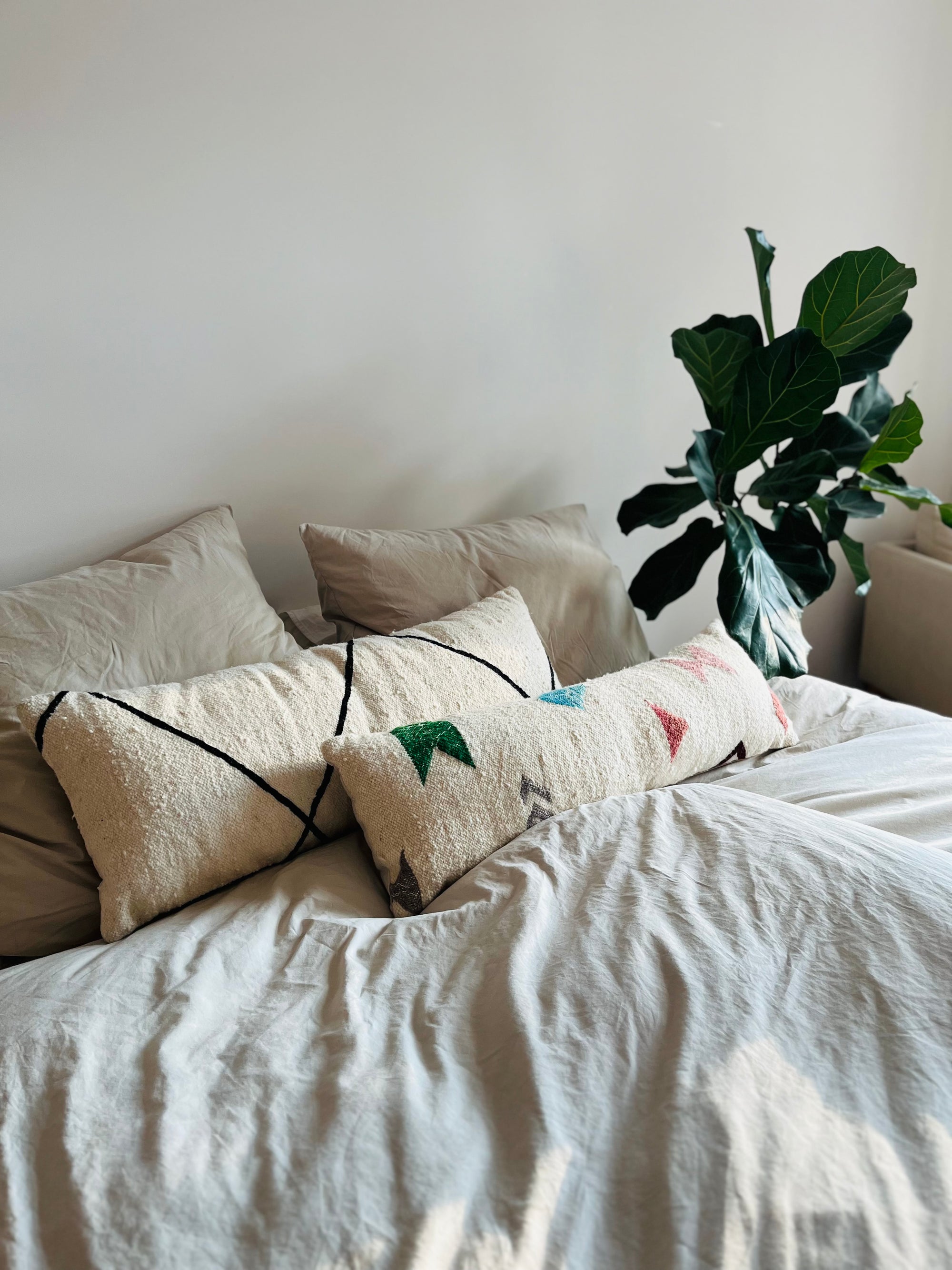 Ein ordentlich gemachtes Bett mit hellbeiger Bettwäsche, zwei LEY! Kissen - Colorful Line - 35 x 90 von LEY! und eine grüne Blattpflanze in der Ecke vor einer schlichten, cremefarbenen Wand schaffen eine ruhige und gemütliche Atmosphäre im Schlafzimmer.