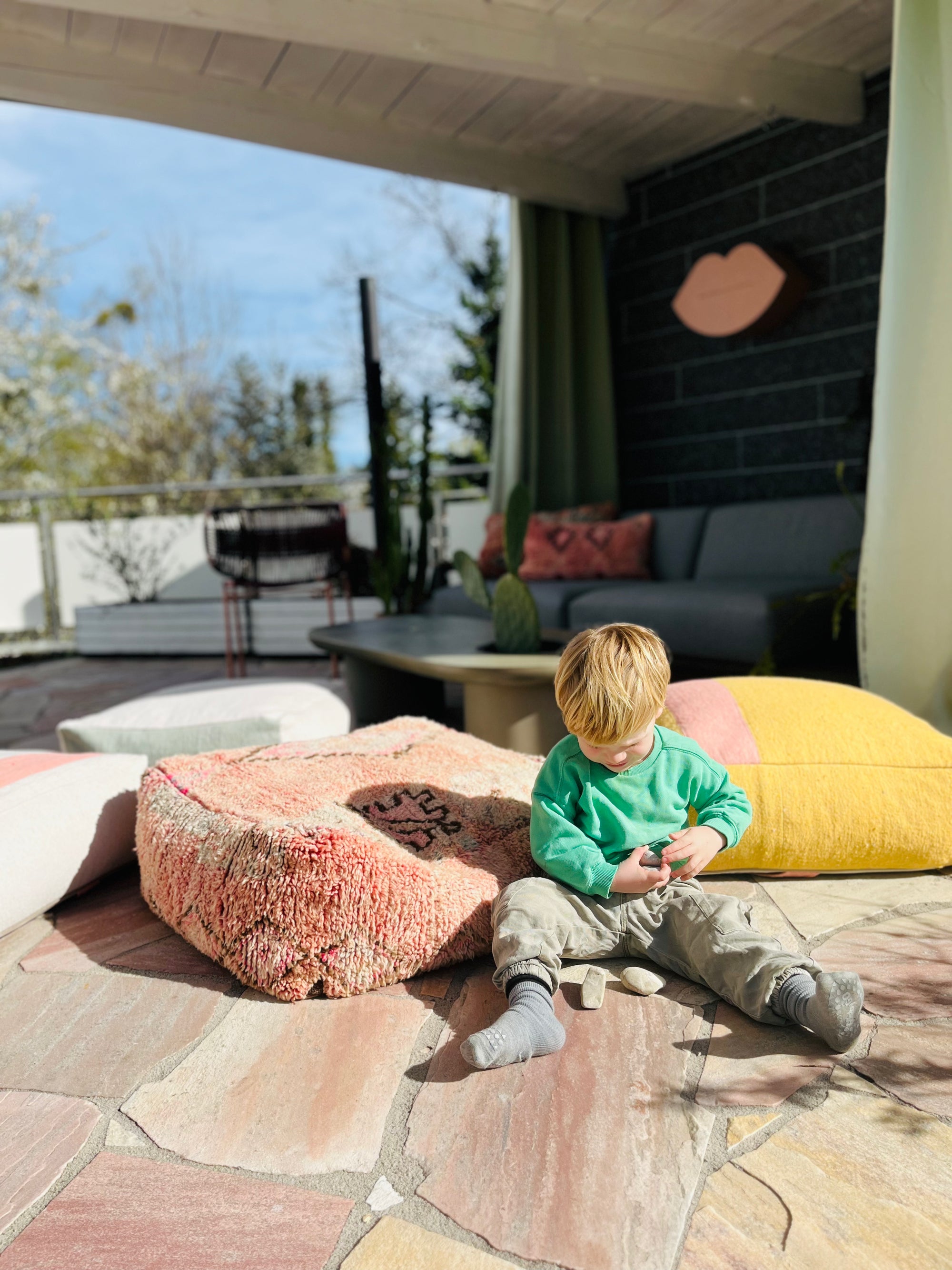 Ein kleines Kind sitzt auf einer Steinterrasse im Sonnenlicht und spielt mit kleinen Steinen. Hinter ihm befinden sich bunte Kissen und ein Sofa, im Hintergrund sind Bäume und ein Zaun zu sehen. Die Szene scheint ruhig und im Freien zu sein.