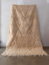 Der BERBER TEPPICH "JULA" von LEY! mit den Maßen 2,00x3,10m ist ein großer, beigefarbener, handgeknüpfter Teppich mit Rautenmuster und kurzen Fransen, der aufrecht vor einer schlichten weißen Wand auf einem glatten Betonboden steht.